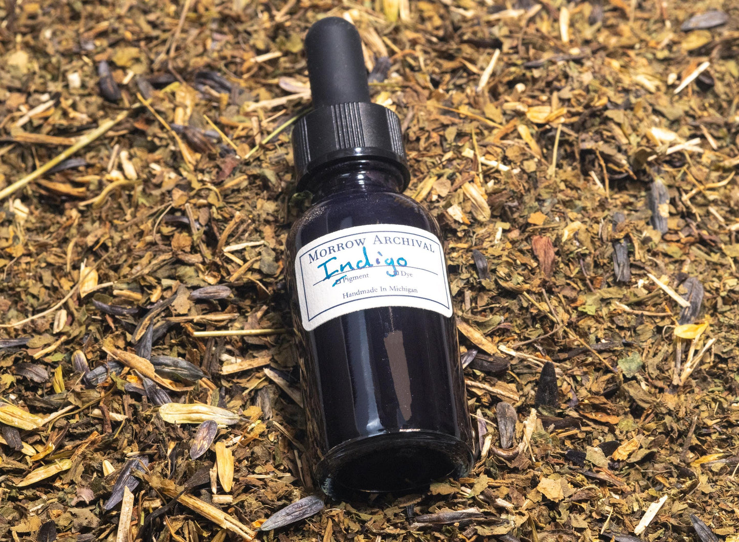 Glass bottle of natural Indigo ink resting on dried Indigofera tinctoria leaves and indigo cakes. The scene connects the finished ink to its botanical source, revealing the deep blue dye’s rich cultural and historical roots.