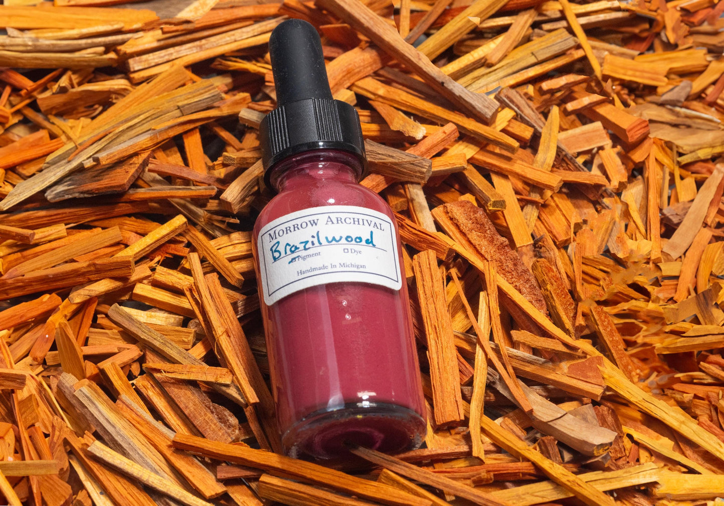Dropper bottle of Brazilwood and Sappanwood Pink Lake ink resting on rough-cut sappanwood chips and shredded brazilwood bark. The raw dye source surrounds the bottle, showcasing the vibrant rose-magenta hue’s origins in natural heartwood.