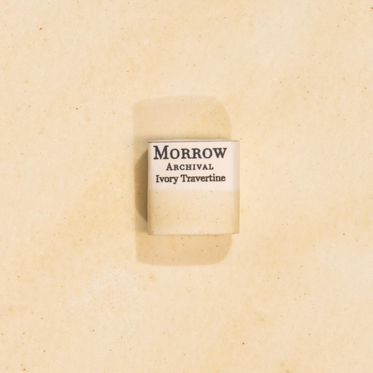 Handmade Ivory Travertine natural watercolor paint half pan resting on a textured watercolor painting. Made from pure single-pigment natural stone, this soft, warm off-white offers subtle granulation and delicate transparency, perfect for botanical painting, historical artwork, and fine art layering. A beautiful eco-friendly gift for watercolor artists and collectors seeking small-batch, archival-quality, all-natural handmade pigments inspired by ancient materials.