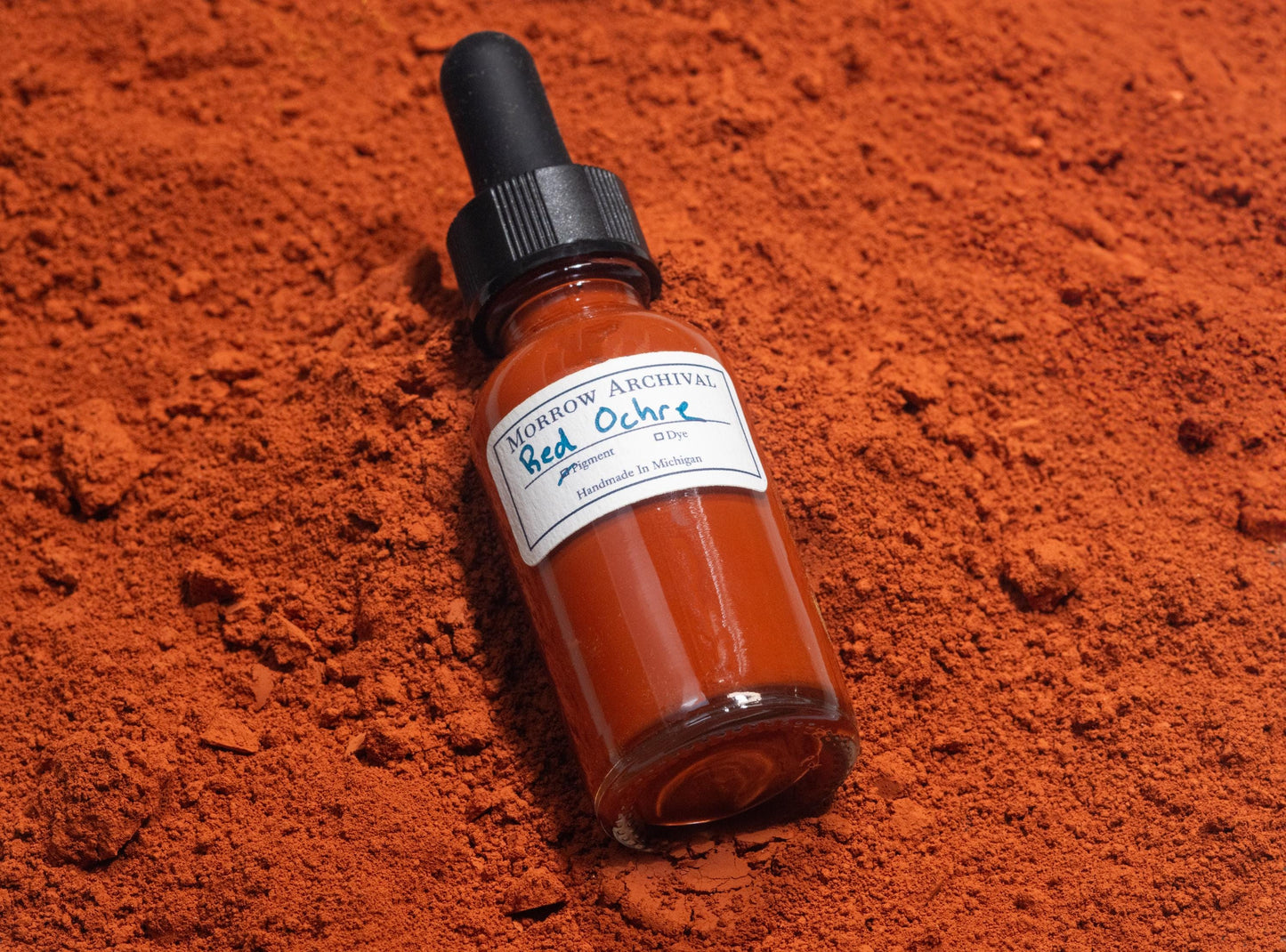 Ink bottle filled with Italian Red Ochre resting on crushed red ochre stones and natural earth samples from Italy. The warm, iron-rich tones of the raw mineral enhance the authenticity of this traditional pigment and its historical applications.