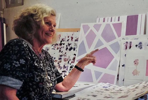 A smiling woman in a floral shirt is pointing at a large abstract painting on a table.