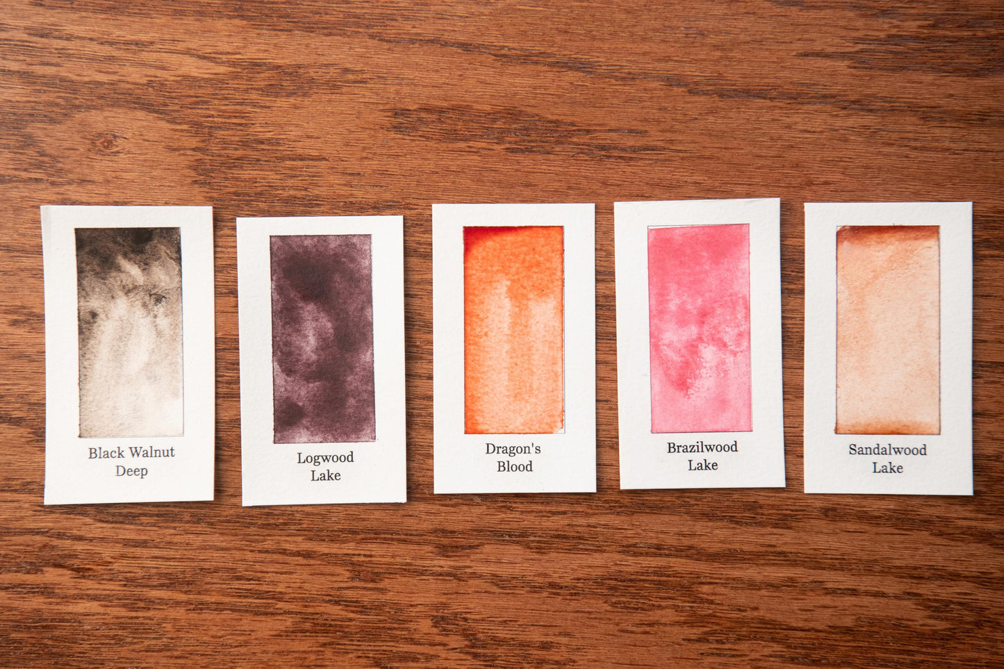 five watercolor paint swatches arranged on a wooden surface, each with a different color and label.