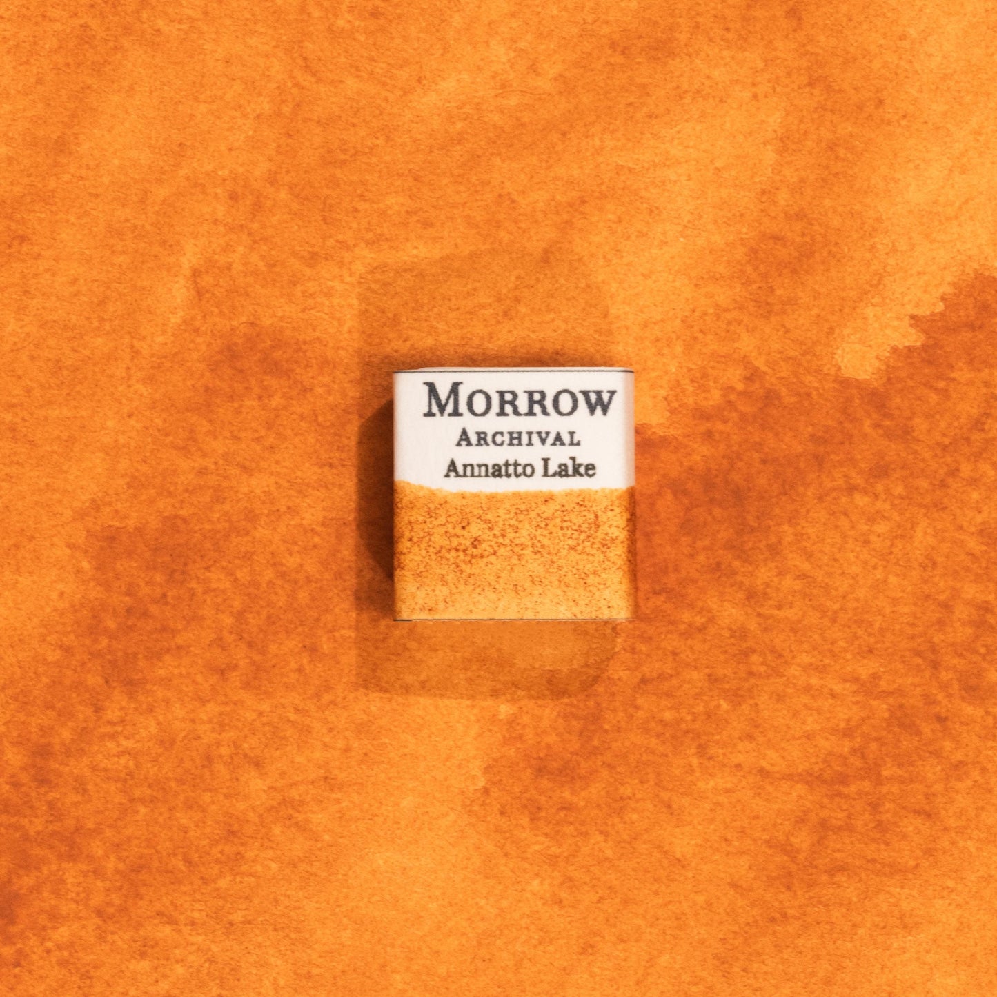 Handmade Annatto Lake Orange natural watercolor paint half pan shown on a textured watercolor painting. Created from pure single-pigment annatto seeds, this warm, glowing orange offers beautiful transparency and soft granulation, ideal for botanical art, historical painting, and fine art illustration. A perfect eco-friendly gift for watercolor artists, nature lovers, and collectors seeking small-batch, archival-quality, all-natural handmade pigments.