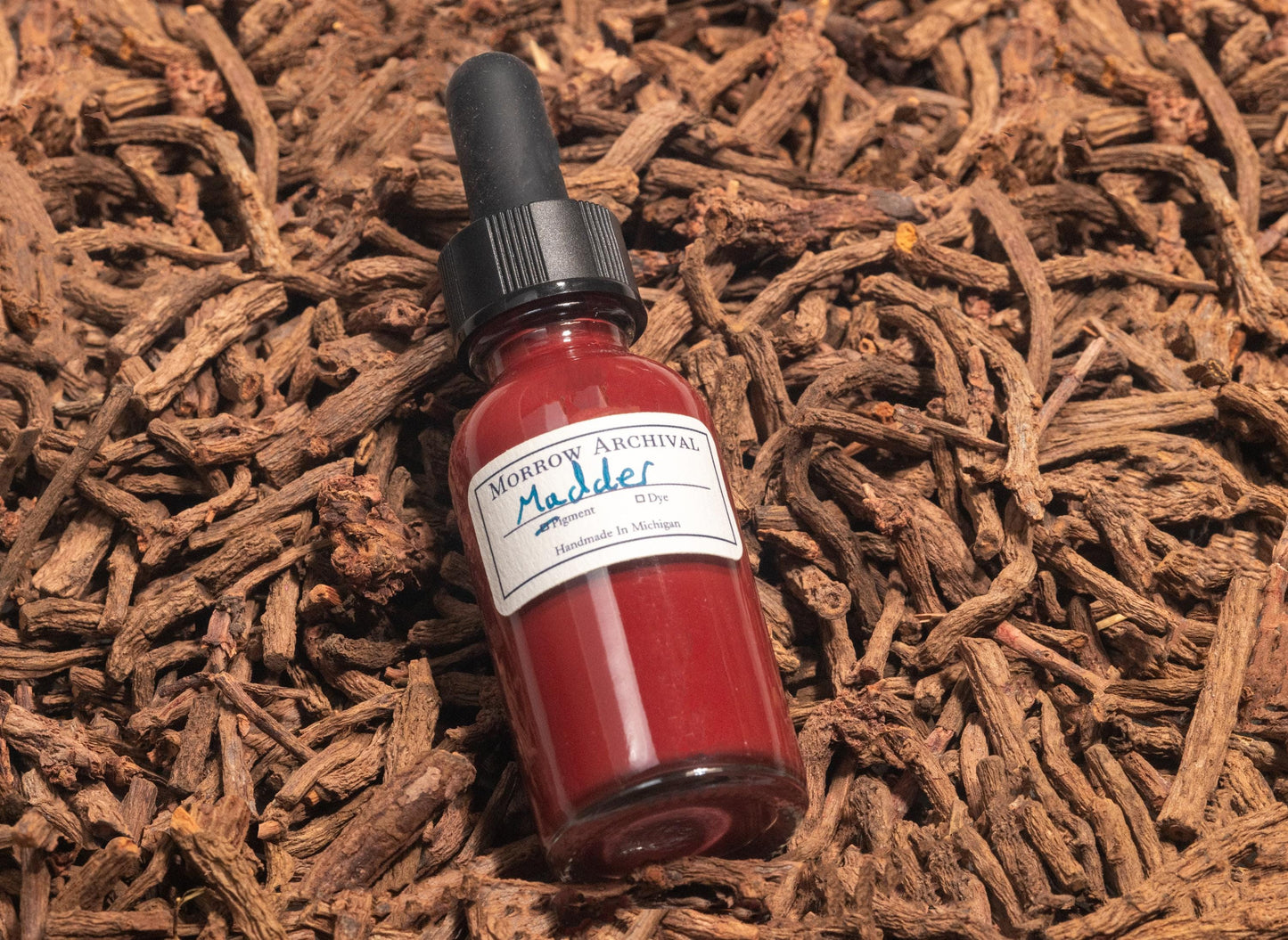 Glass bottle of Madder Red Lake ink resting on dried and fermented Rubia tinctorum roots. The image showcases the warm red ink’s botanical origin and deep ties to historical European and Middle Eastern textile and art traditions.
