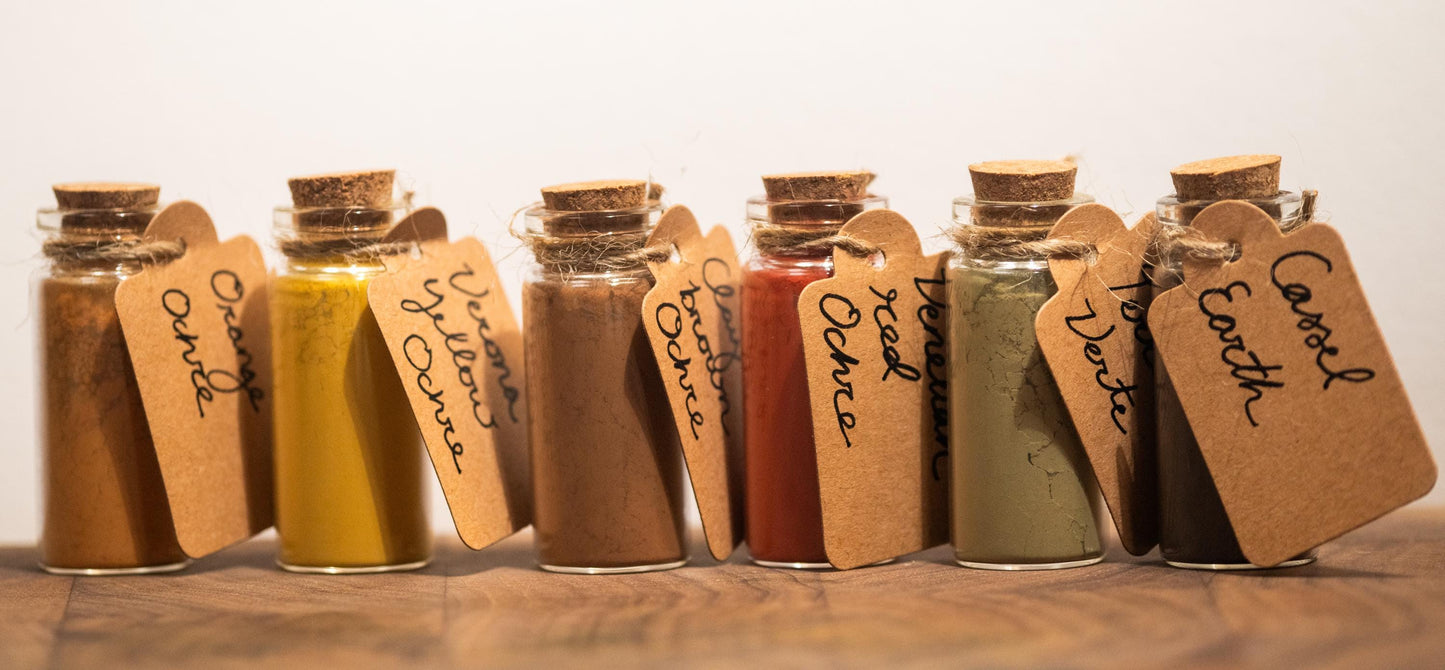 Seven pigment bottles removed from the box and arranged in a neat line, each labeled in hand-calligraphed tags identifying the ochres: Verona Yellow, Red, Orange, Clay Brown, Venetian Red, Terre Verte, and Cassel Earth. The bottles are displayed with care and elegance, emphasizing the artisanal, natural qualities of each earthy pigment.