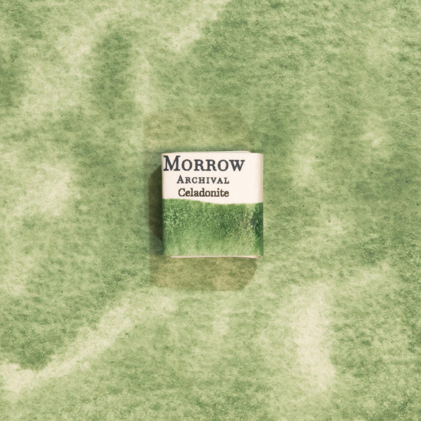 Handmade Celadonite Green Earth ochre natural watercolor paint half pan resting on a textured watercolor painting. Crafted from pure single-pigment celadonite mineral, this soft, muted green offers beautiful transparency and fine granulation, ideal for landscape painting, botanical illustration, and historical artwork. A perfect eco-friendly gift for watercolor artists and collectors seeking small-batch, archival-quality, all-natural handmade pigments with ancient roots.