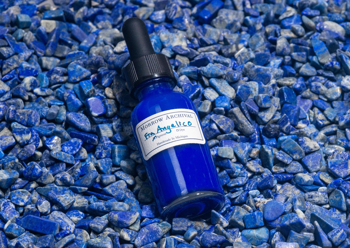 Clear glass bottle of Fra Angelico Blue ink nestled on raw lapis lazuli stones, showing the mineral’s ultramarine blue veins. The iconic semi-precious stone underscores the historic and luxurious origin of this deep celestial pigment.