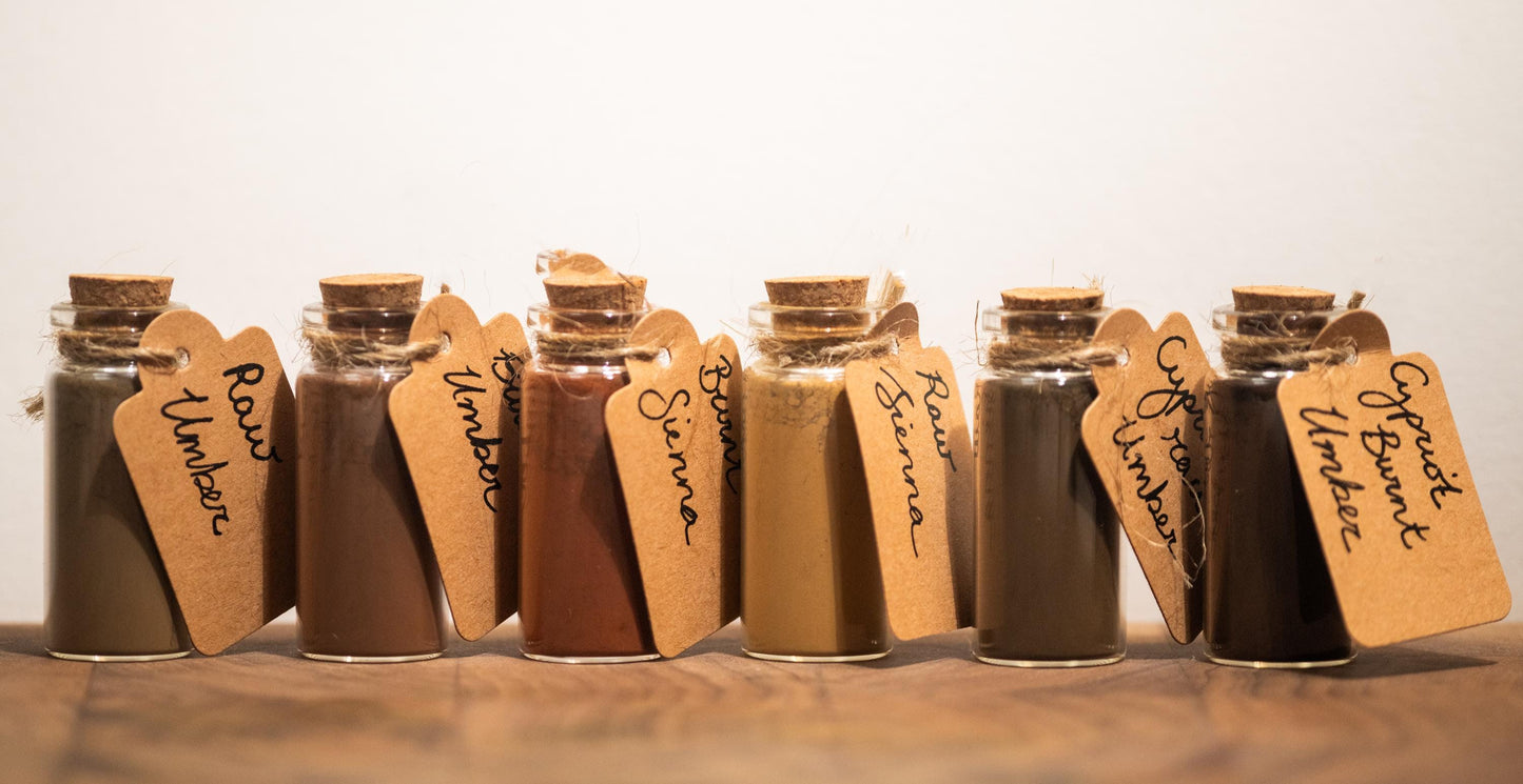 Six labeled pigment bottles arranged in an artful display with handwritten calligraphy tags reading Raw Umber, Burnt Umber, Raw Sienna, Burnt Sienna, Cypriot Raw Umber, and Cypriot Burnt Umber. This photo showcases the natural variation in tone and character of each pigment, emphasizing their use in timeless palettes.