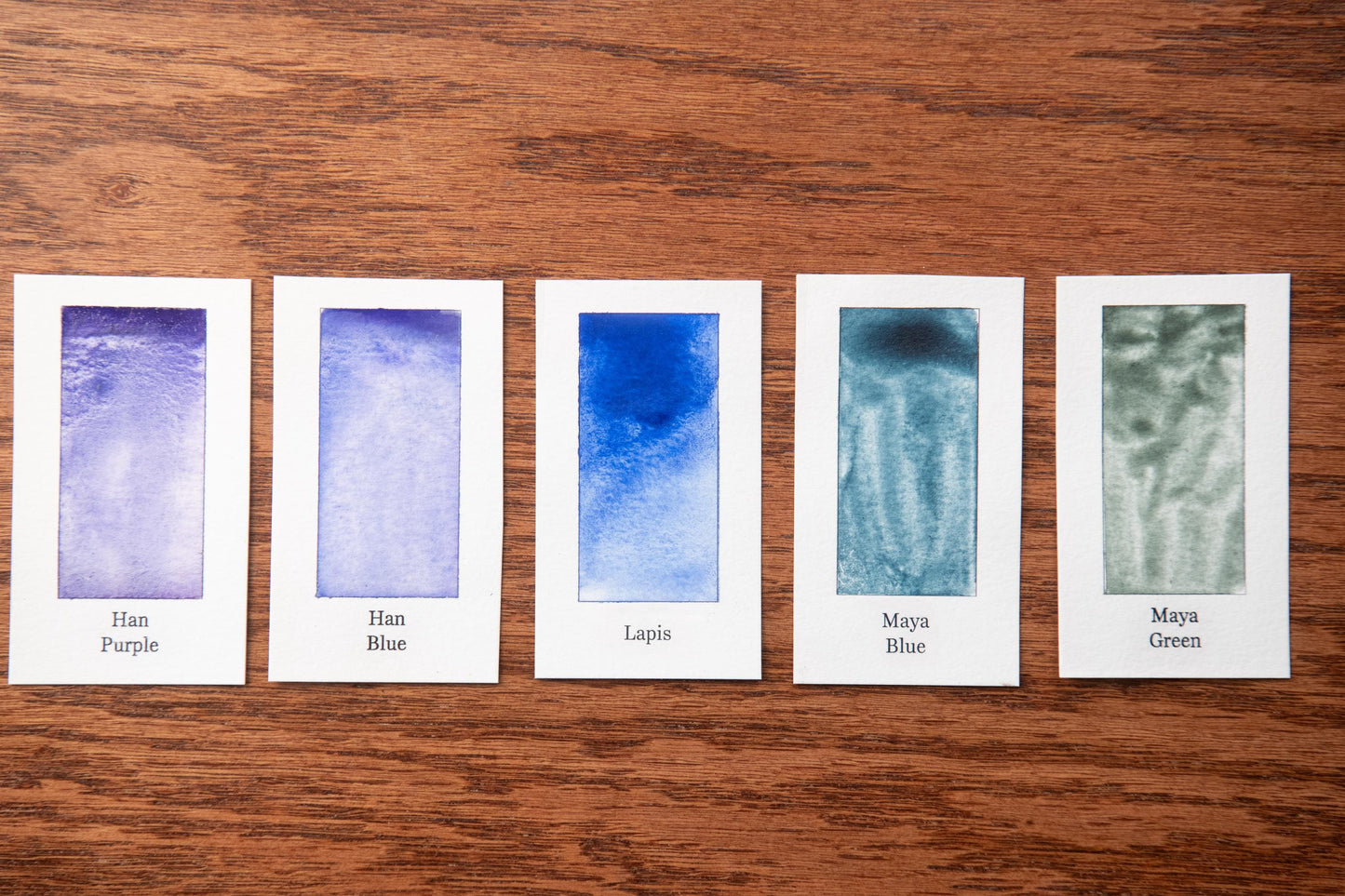 five watercolor paintings with different shades of blue, arranged on a wooden surface.