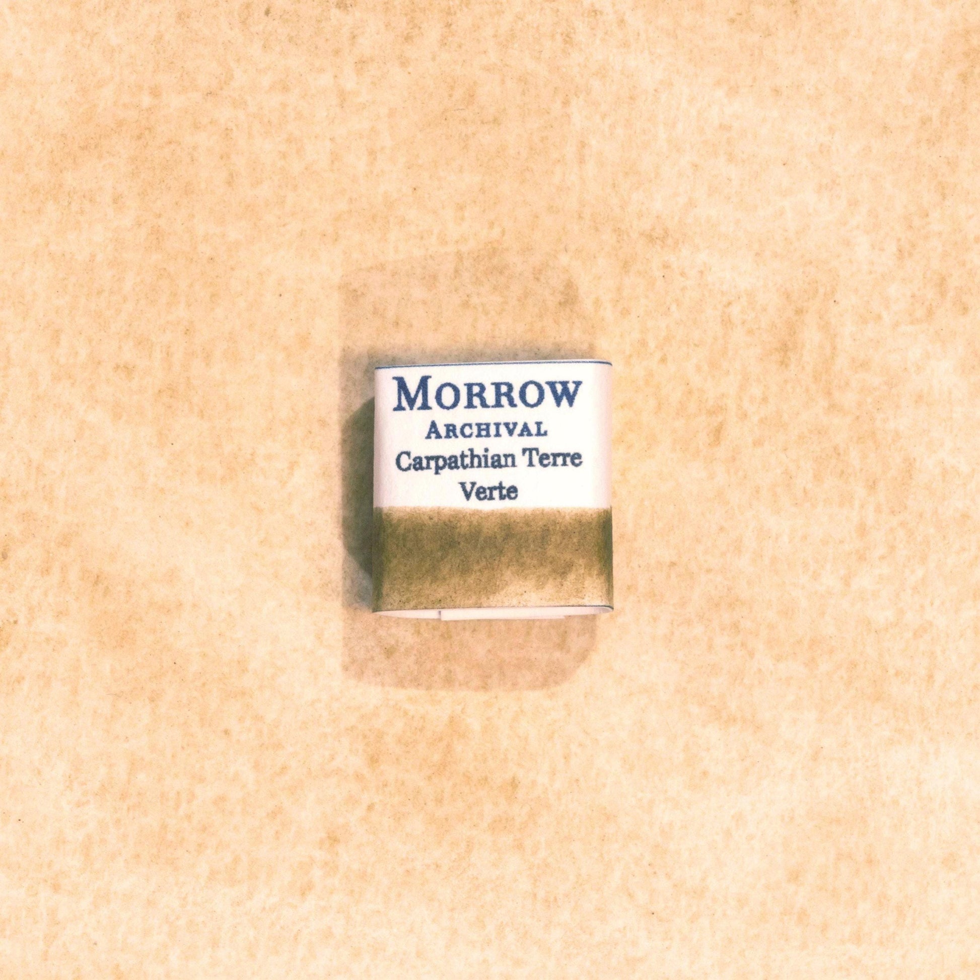 Handmade Carpathian Terre Verte natural watercolor paint half pan displayed on a textured watercolor painting. Crafted from pure single-pigment green earth sourced from the Carpathian Mountains, offering soft, muted green tones perfect for classical art, botanical work, and landscape painting. An ideal eco-friendly gift for artists, watercolorists, and historians who appreciate archival-quality, small-batch, all-natural pigments.