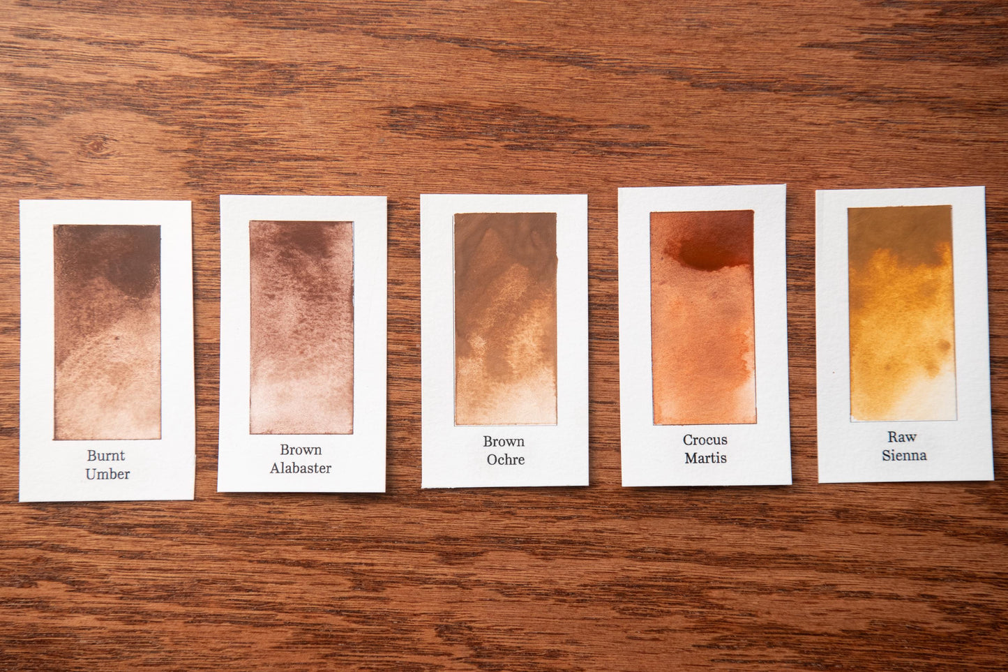 five small rectangular cards with different shades of brown paint, arranged on a wooden surface.