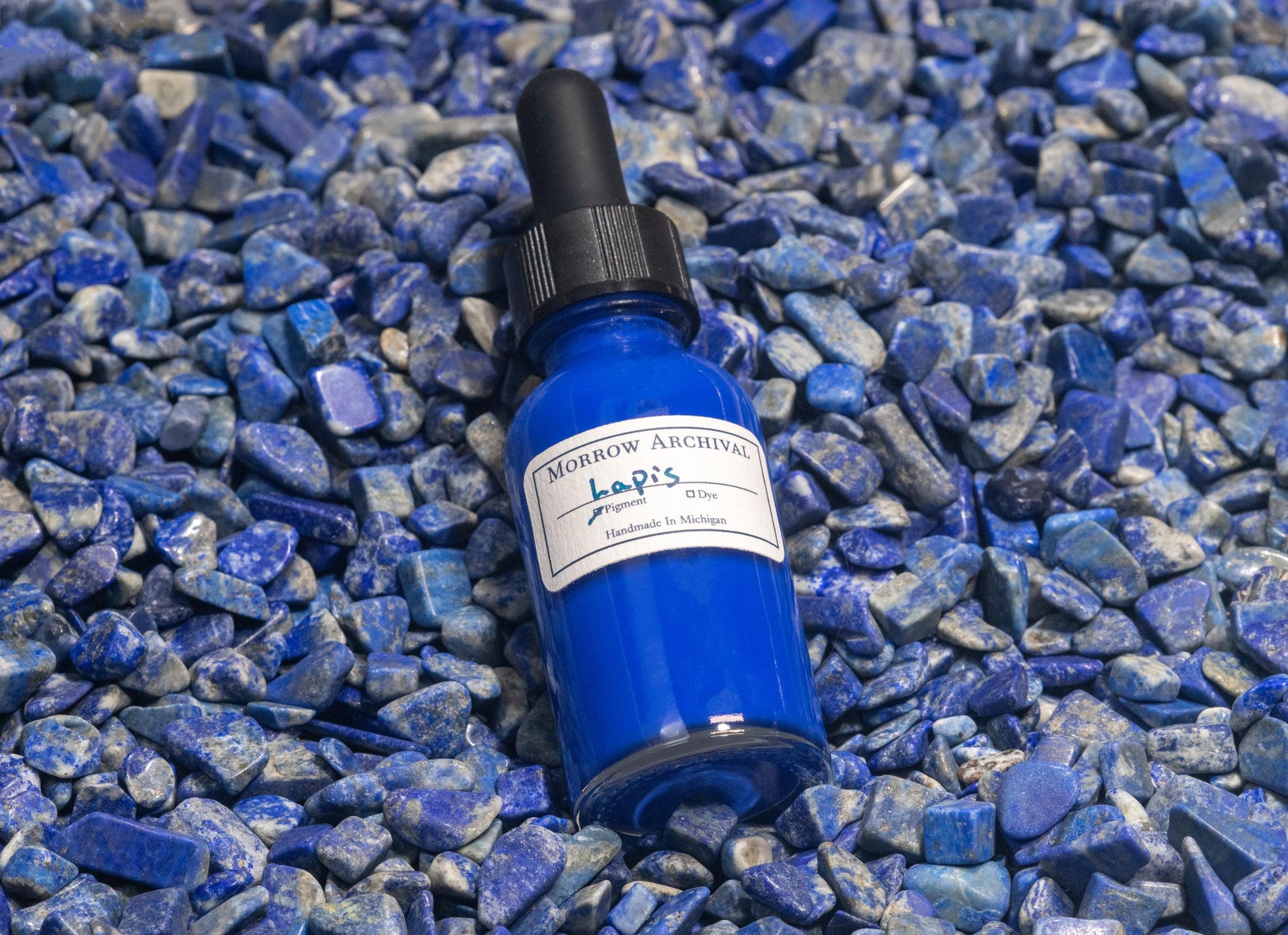 Natural Lapis Lazuli ink bottle resting on ground lapis pigment and uncut stone fragments. The setup links the glowing ultramarine ink to its semi-precious origin, emphasizing its mineral brilliance and historical prestige.