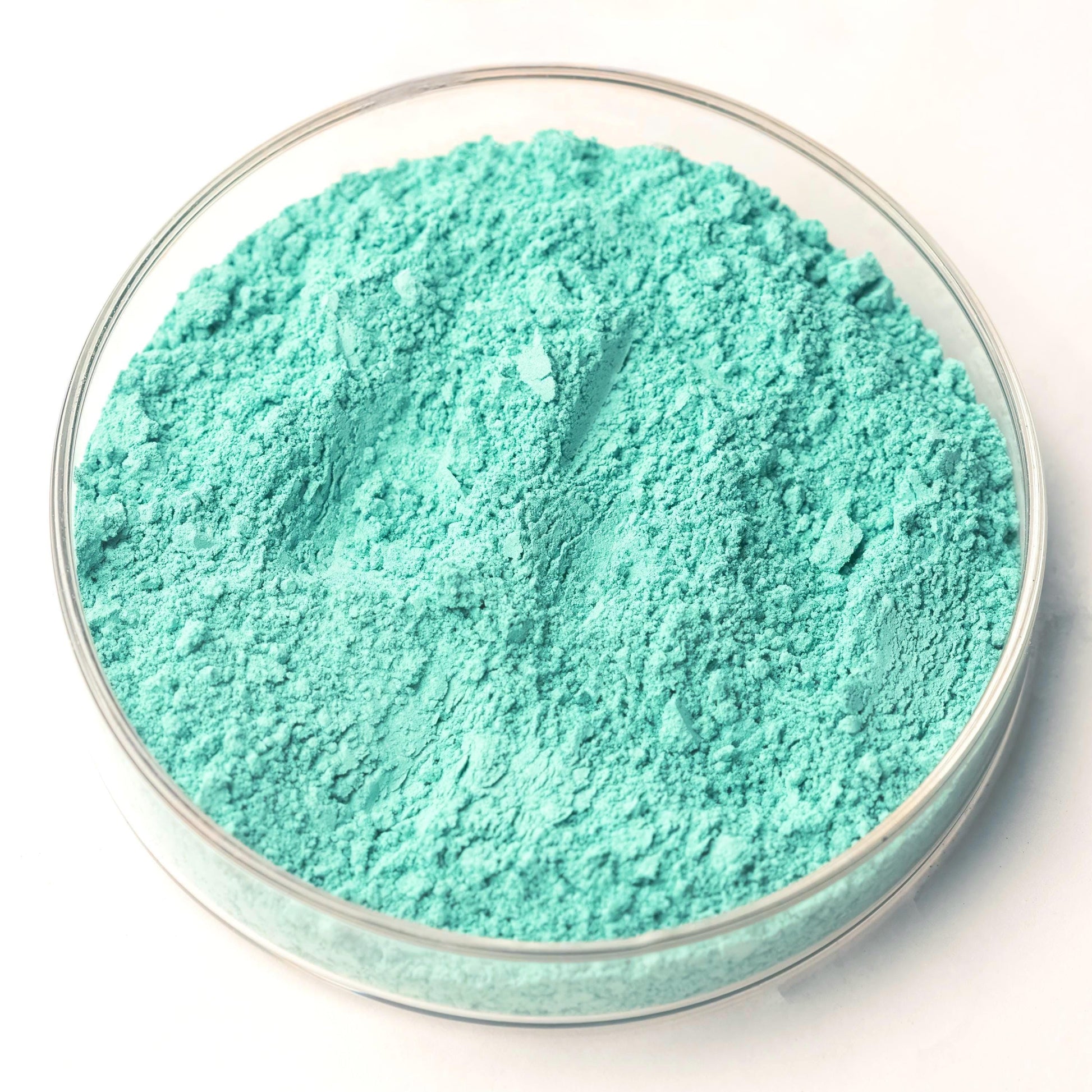 A clear glass dish containing Green Verditer Pigment, a bright turquoise blue-green powder, sits elegantly on a white surface.