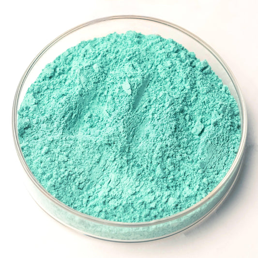 A clear glass dish containing Green Verditer Pigment, a bright turquoise blue-green powder, sits elegantly on a white surface.