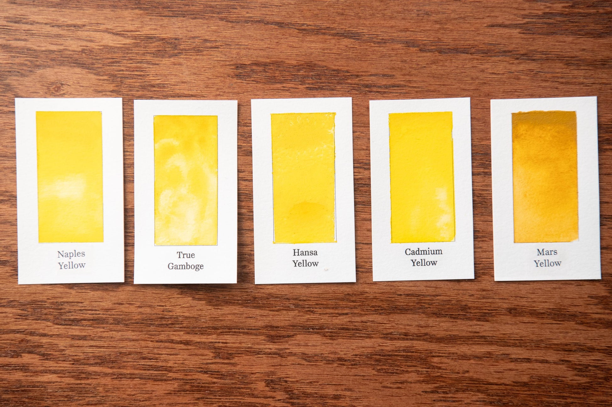 five identical cards, each containing a different shade of yellow paint. The cards are arranged in a row on a wooden surface, with each card labeled with a different shade of yellow paint.