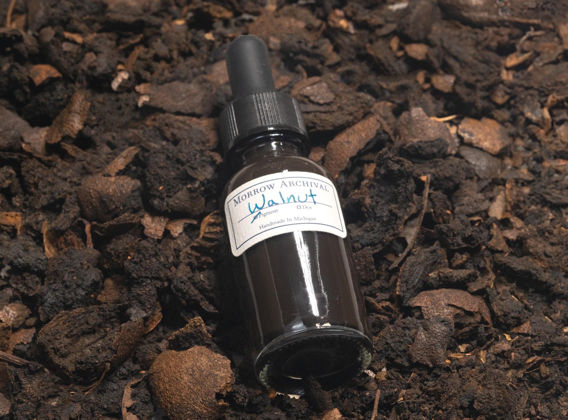 Black Walnut Lake ink bottle sitting on a bed of cracked walnut hulls and powdered dye. The dark brown hues of the husks highlight the natural botanical source of this warm, transparent ink used in historical American and European traditions.