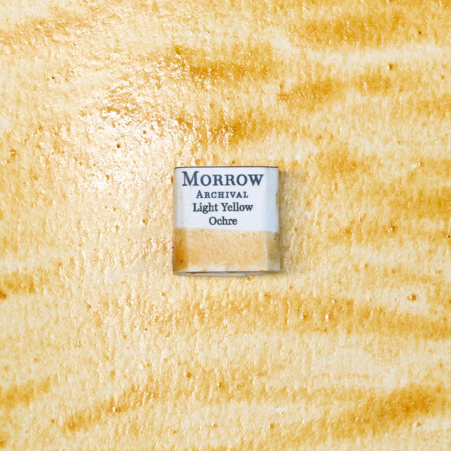 Close-up image of a half pan of Light Yellow Ochre handmade watercolor paint, showing a soft, warm golden-yellow color with a smooth, matte texture. The pan rests on a neutral-toned background, highlighting its natural earth origin and artisanal, small-batch craftsmanship. Ideal for artists seeking a timeless, light ochre pigment with historical significance and rich, natural tone.