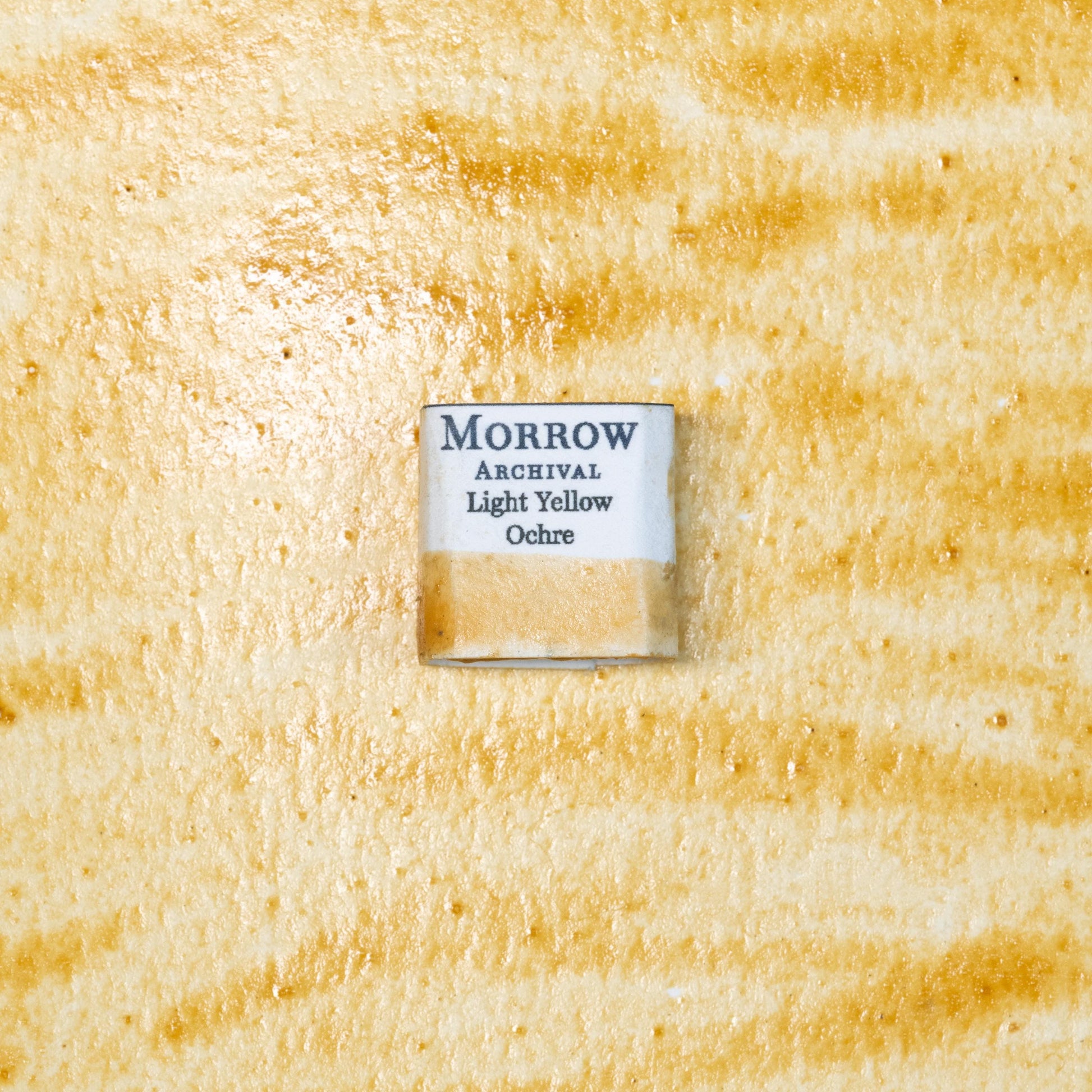 Close-up image of a half pan of Light Yellow Ochre handmade watercolor paint, showing a soft, warm golden-yellow color with a smooth, matte texture. The pan rests on a neutral-toned background, highlighting its natural earth origin and artisanal, small-batch craftsmanship. Ideal for artists seeking a timeless, light ochre pigment with historical significance and rich, natural tone.
