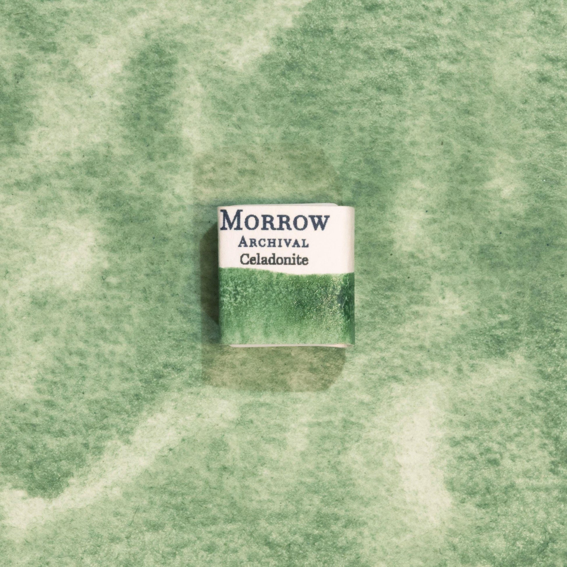 Handmade Celadonite Green Earth ochre natural watercolor paint half pan resting on a textured watercolor painting. Crafted from pure single-pigment celadonite mineral, this soft, muted green offers beautiful transparency and fine granulation, ideal for landscape painting, botanical illustration, and historical artwork. A perfect eco-friendly gift for watercolor artists and collectors seeking small-batch, archival-quality, all-natural handmade pigments with ancient roots.