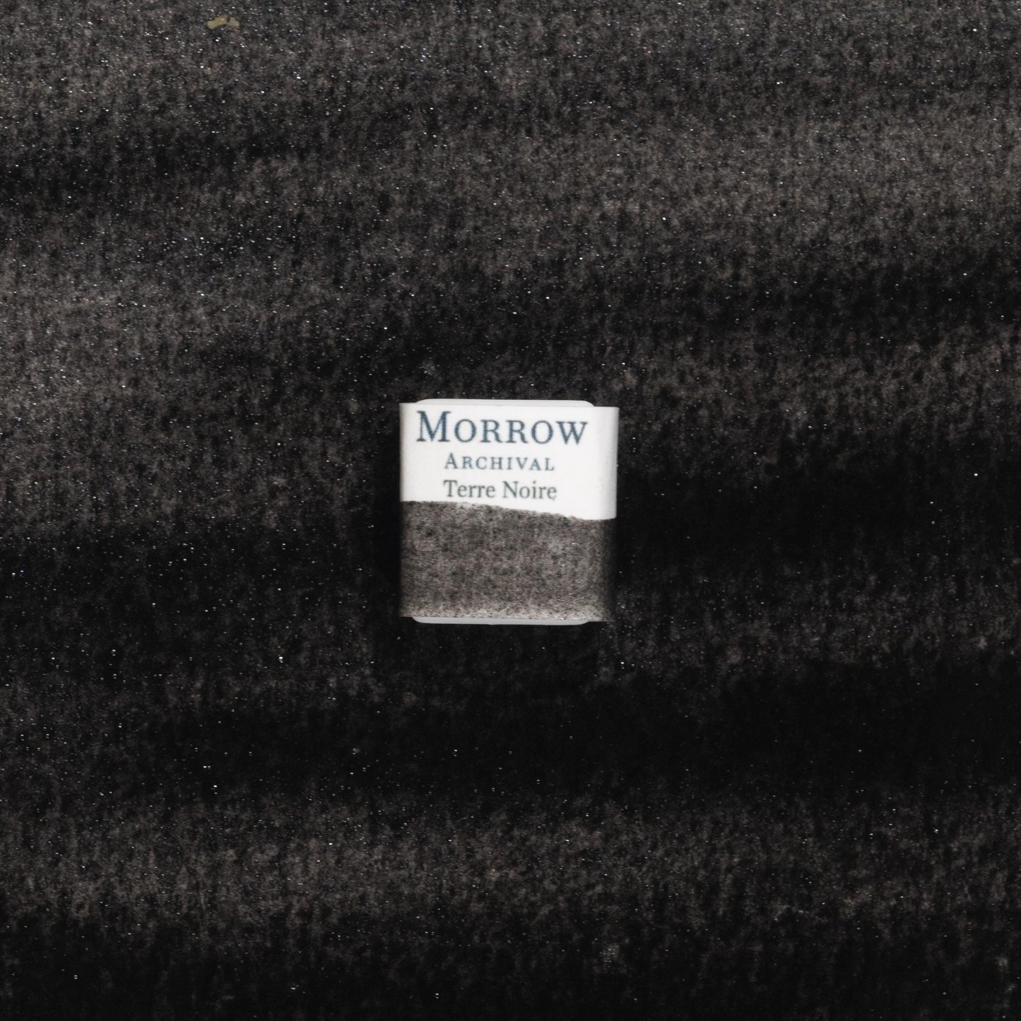 Handmade Terre Noire Black Earth natural watercolor paint half pan resting on a textured watercolor painting. Crafted from pure single-pigment black earth, this deep, matte black offers subtle granulation and natural undertones, ideal for landscape shadows, historical painting, and monochrome studies. A bold eco-friendly gift for watercolor artists and collectors seeking small-batch, archival-quality, all-natural handmade pigments rooted in ancient earth traditions.