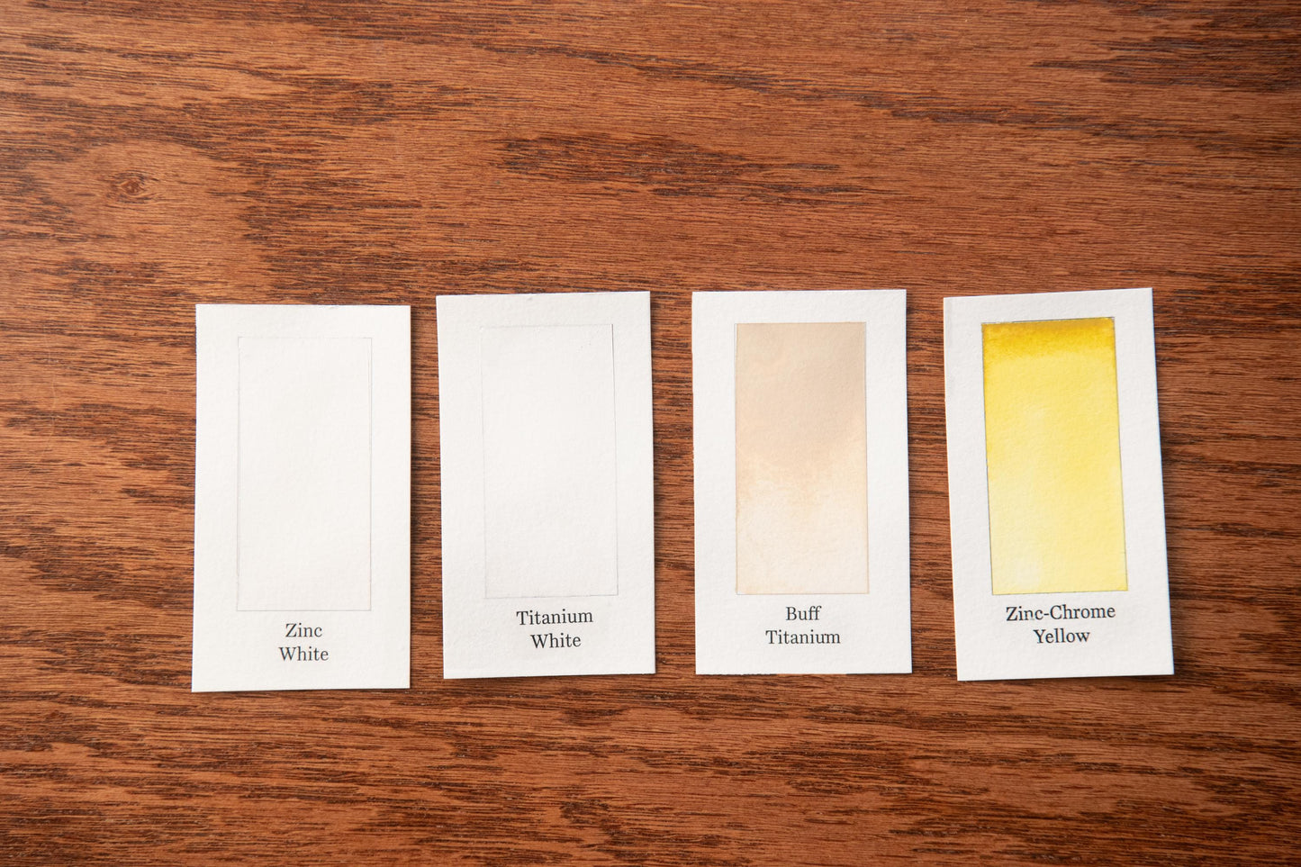 four color swatches on a wooden surface, each representing a different color: white, silver, gold, and yellow.