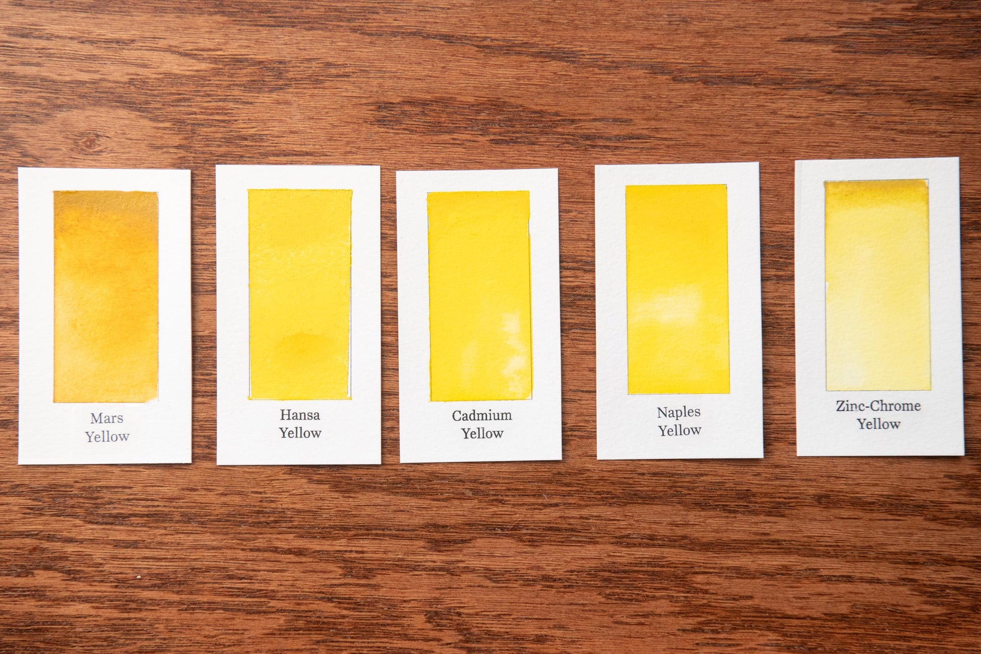 five identical cards, each containing a different shade of yellow paint. The cards are arranged in a row on a wooden surface, with each card labeled with the name of the yellow paint shade it represents.