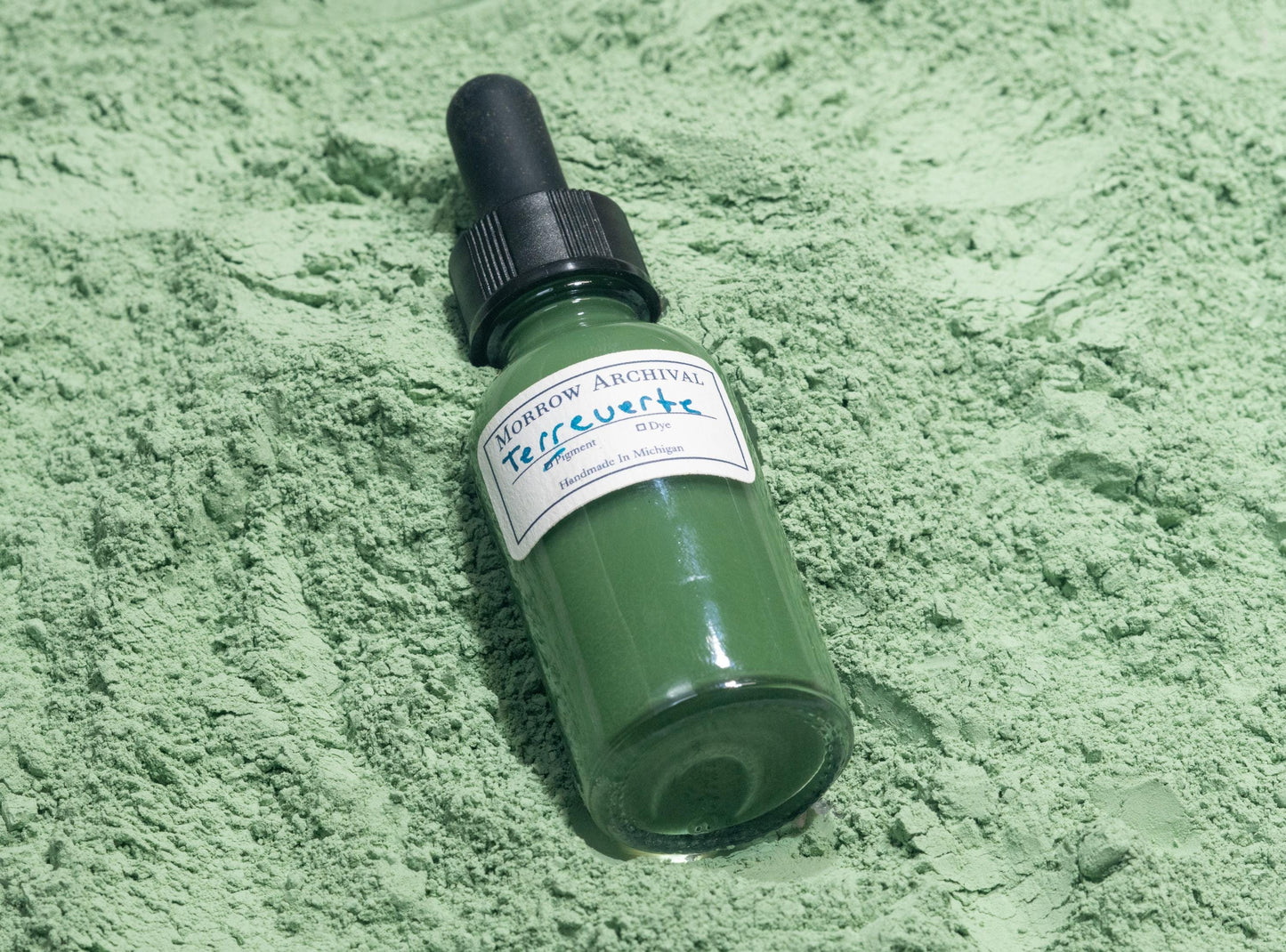 Bottle of Terre Verte ink placed on natural green earth powder and clay nodules. The subdued sage and olive hues of the raw mineral reflect the gentle tone and long artistic history of this pigment in underpaintings and figure work.