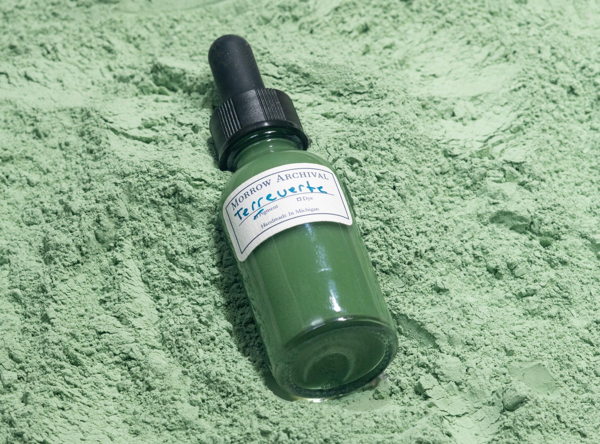 Bottle of Terre Verte ink placed on natural green earth powder and clay nodules. The subdued sage and olive hues of the raw mineral reflect the gentle tone and long artistic history of this pigment in underpaintings and figure work.