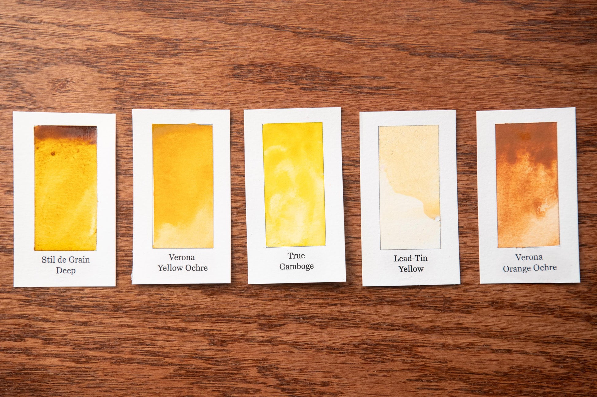five small square cards with different shades of yellow paint, arranged on a wooden surface.