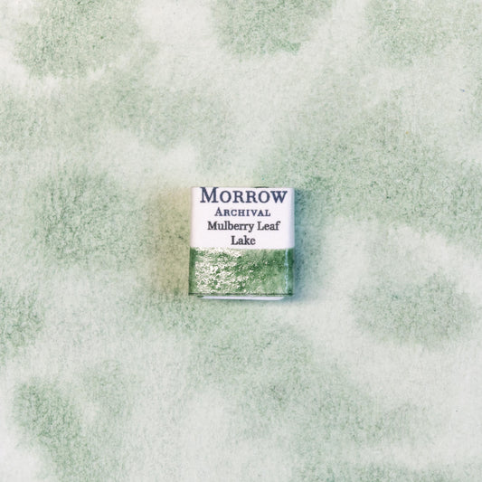 Close-up image of a half pan of Mulberry Leaf Green handmade watercolor paint, showing a soft, natural green with golden undertones and a translucent finish. The pan is placed on a neutral or botanical-inspired background, highlighting its plant-based origin and artisanal craftsmanship. Ideal for artists seeking a delicate, sustainable green with a living, organic feel.
