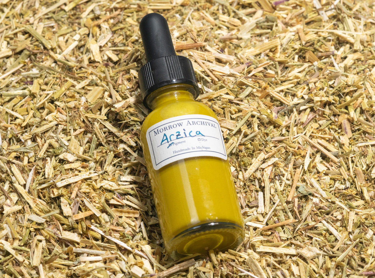 Glass bottle of Arzica Weld Lake ink resting atop dried cut weld stems and flowers, the botanical source of its bright lemon-yellow pigment. The scene highlights the historical process of turning natural plant dye into a vivid watercolor ink, with earthy textures and archival presentation.