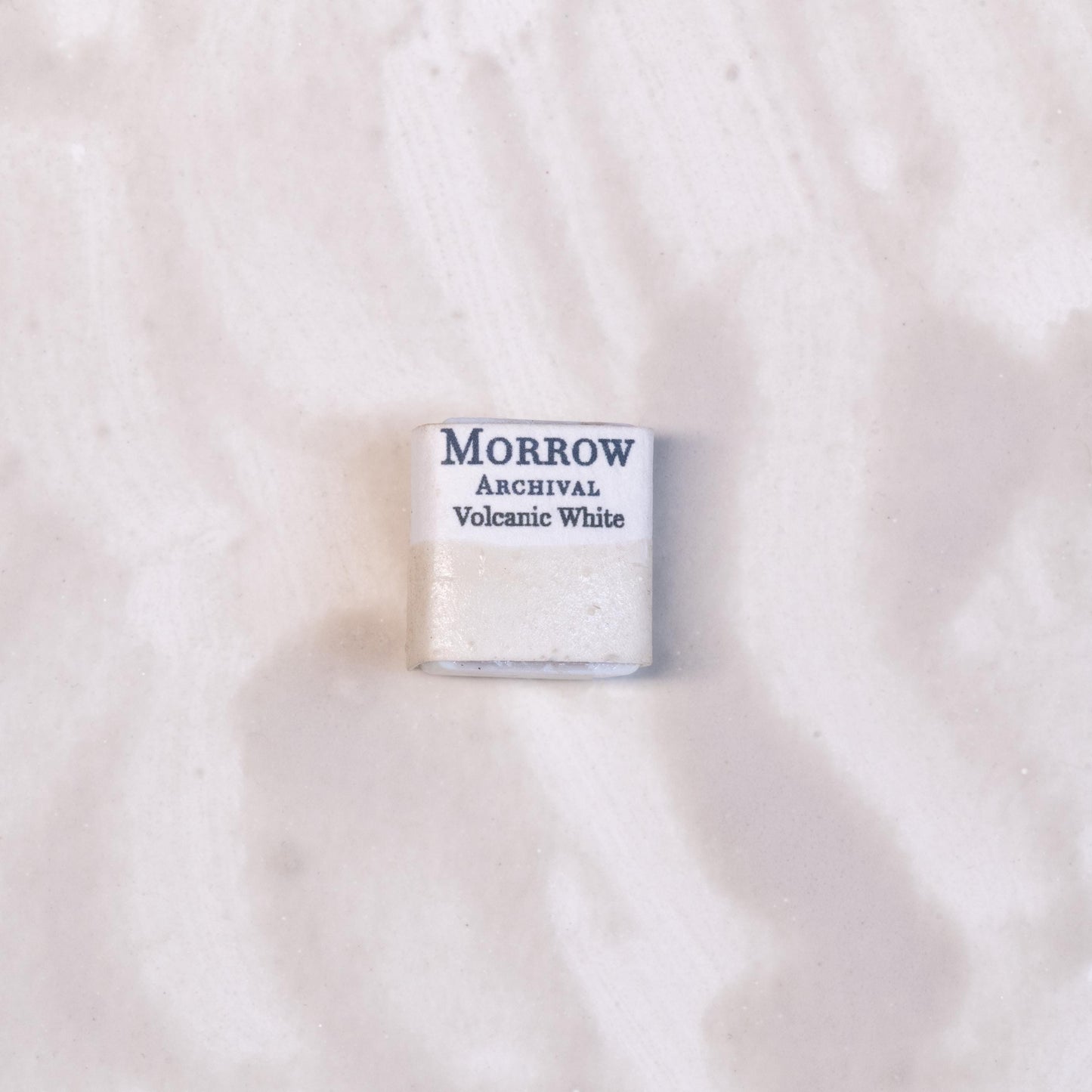 Close-up image of a half pan of Volcanic White handmade watercolor paint, featuring a soft, warm off-white hue with visible natural granulation. The pan is set on a neutral or stone-textured background, highlighting its mineral origin and artisanal craftsmanship. Ideal for artists seeking a textured, natural white with geological roots.