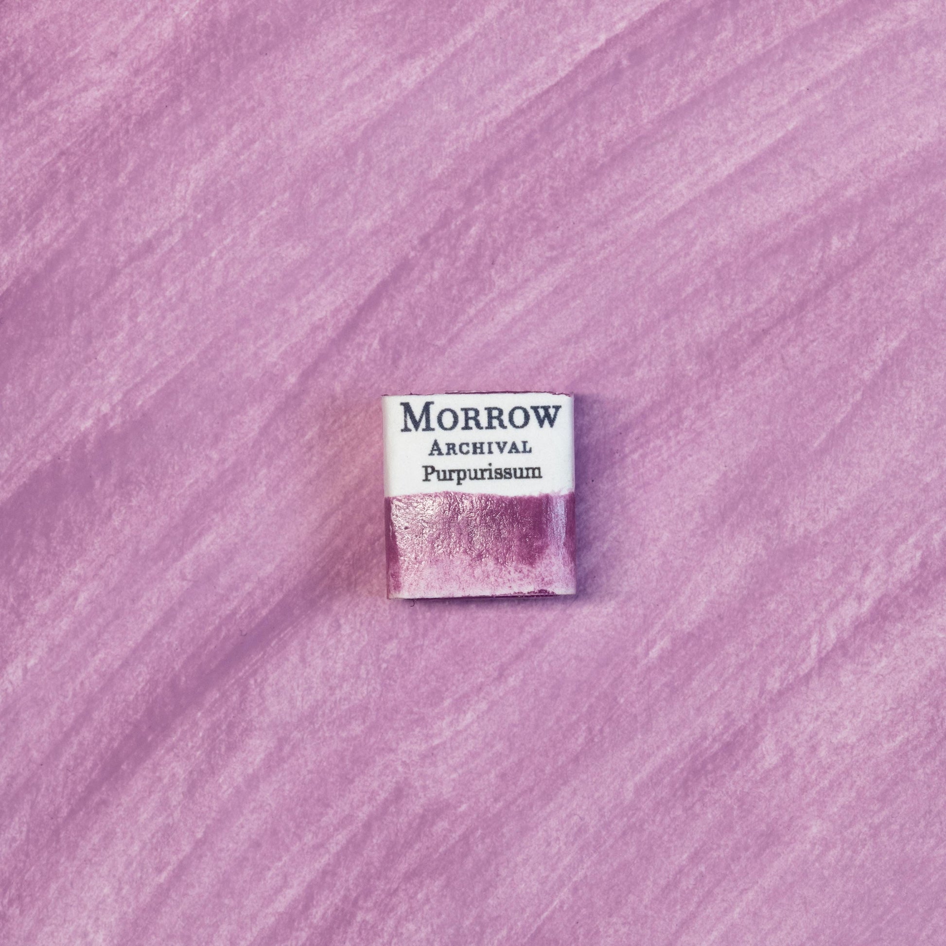 Close-up image of a half pan of Purpurissum handmade watercolor paint, featuring a deep, reddish-purple hue with a smooth, translucent finish and subtle texture. The pan is set on a neutral or ancient-inspired background, emphasizing its origin from Murex sea snails and artisanal craftsmanship. Ideal for artists, collectors, and conservators seeking a true historical pigment of imperial prestige.