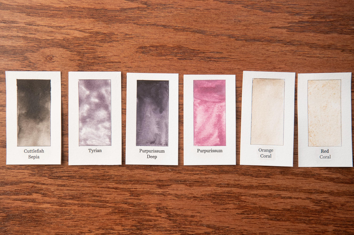 six different watercolor paint swatches arranged on a wooden surface.