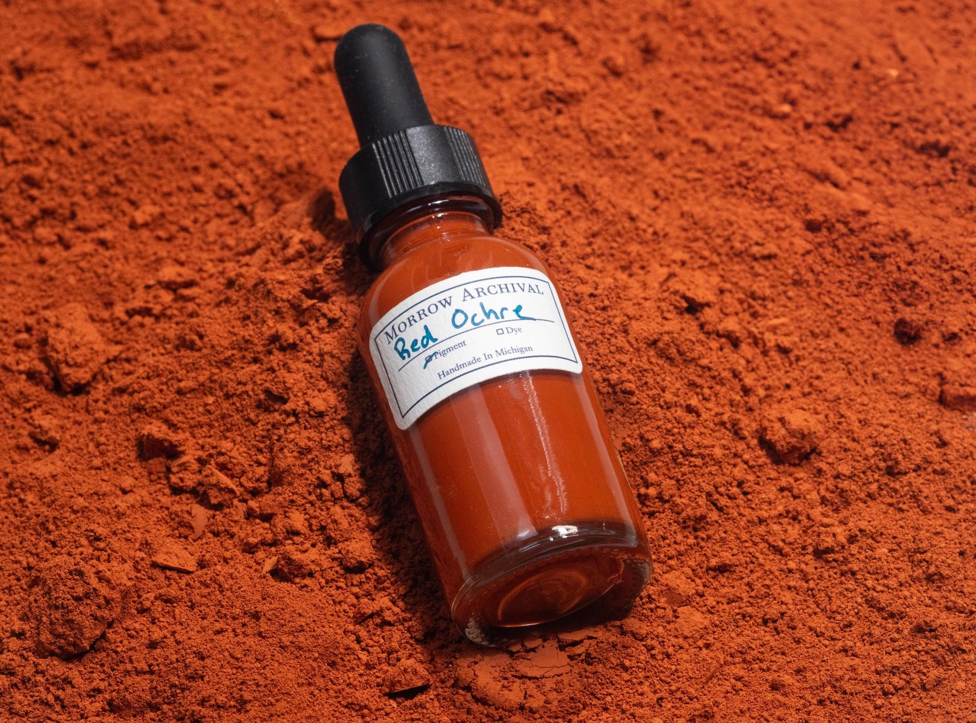 Ink bottle filled with Italian Red Ochre resting on crushed red ochre stones and natural earth samples from Italy. The warm, iron-rich tones of the raw mineral enhance the authenticity of this traditional pigment and its historical applications.