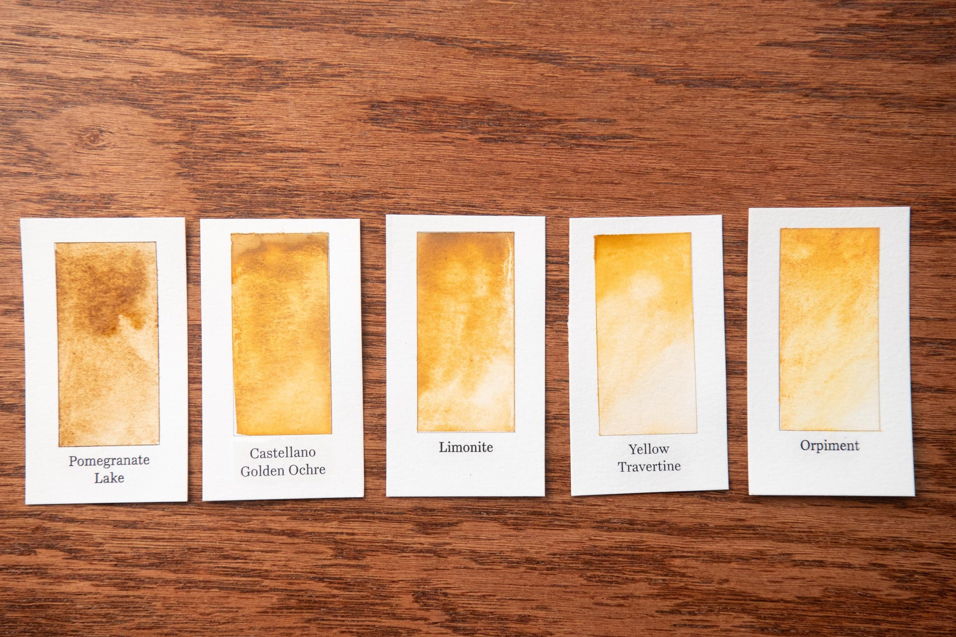 five small cards with different shades of yellow paint, arranged on a wooden surface.