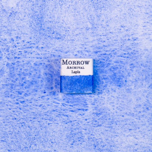 Close-up image of a half pan of Lapis Lazuli handmade watercolor paint, showing a soft, radiant blue with delicate granulation and a translucent matte finish. The pan rests on a neutral background, highlighting its semi-precious stone origin and hand-crafted artistry. Ideal for artists seeking a historically accurate, naturally sourced blue with a luminous, ethereal quality.