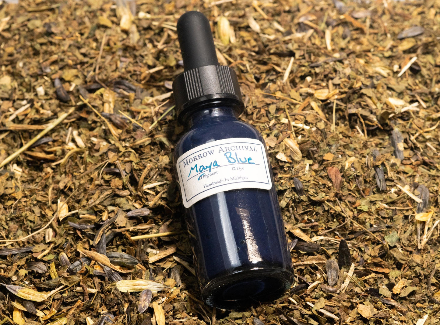 Bottle of True Maya Blue ink sitting on a mix of dried Indigofera suffruticosa leaves and pale palygorskite clay. This composition visually explains the unique Mesoamerican fusion of plant dye and mineral substrate used in this ancient pigment.