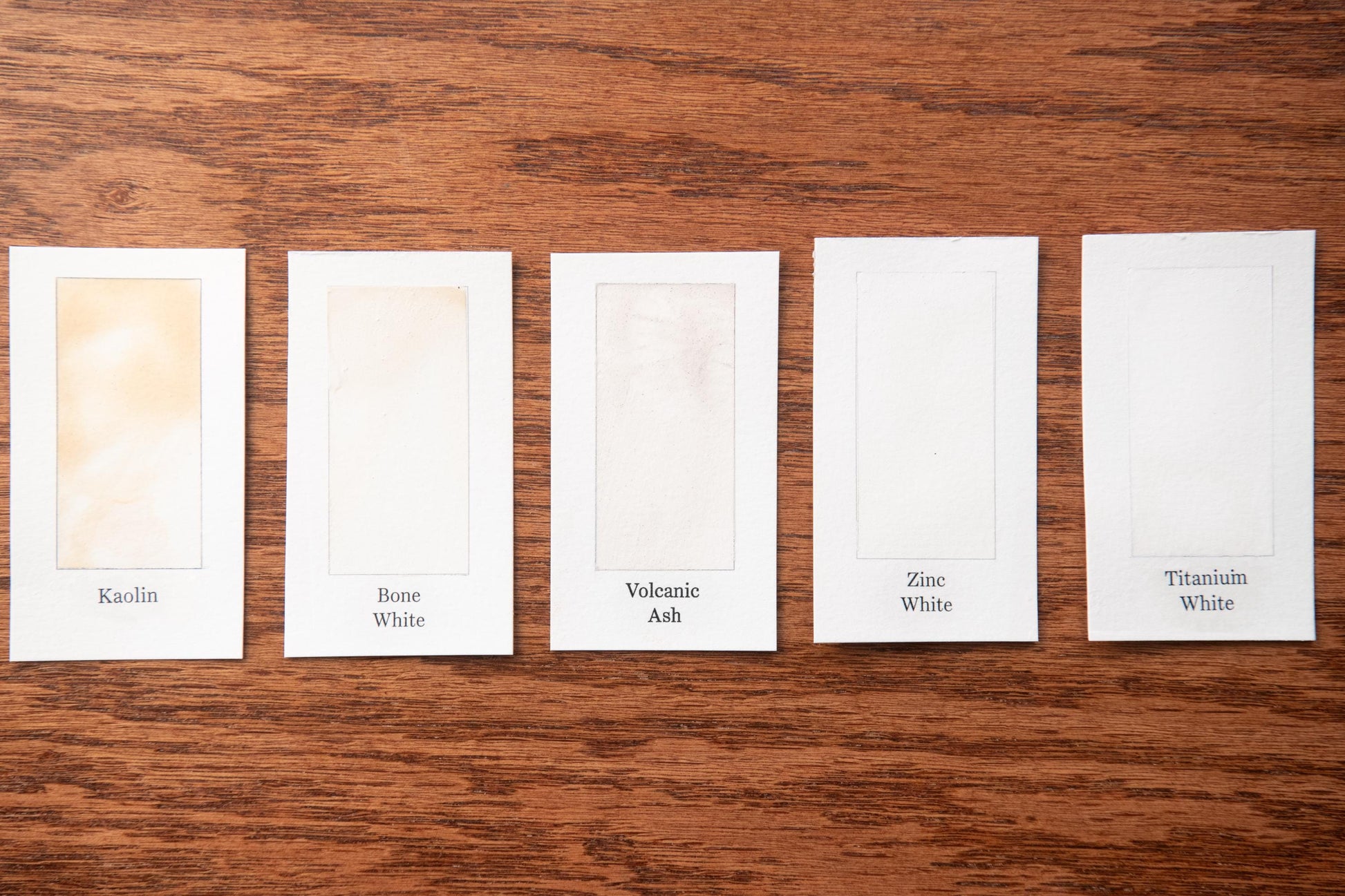 five white cards with different paint colors labeled on them, arranged on a wooden surface.