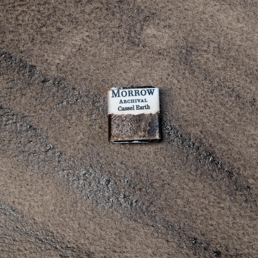 Close-up image of a half pan of Cassel Earth handmade watercolor paint, showing a dark, cool brown with soft granulation and a smooth, semi-transparent matte finish. The pan rests on a neutral or rustic background, emphasizing its historical origin and artisanal craftsmanship. Ideal for artists seeking a moody, expressive brown pigment with classical roots and natural depth.