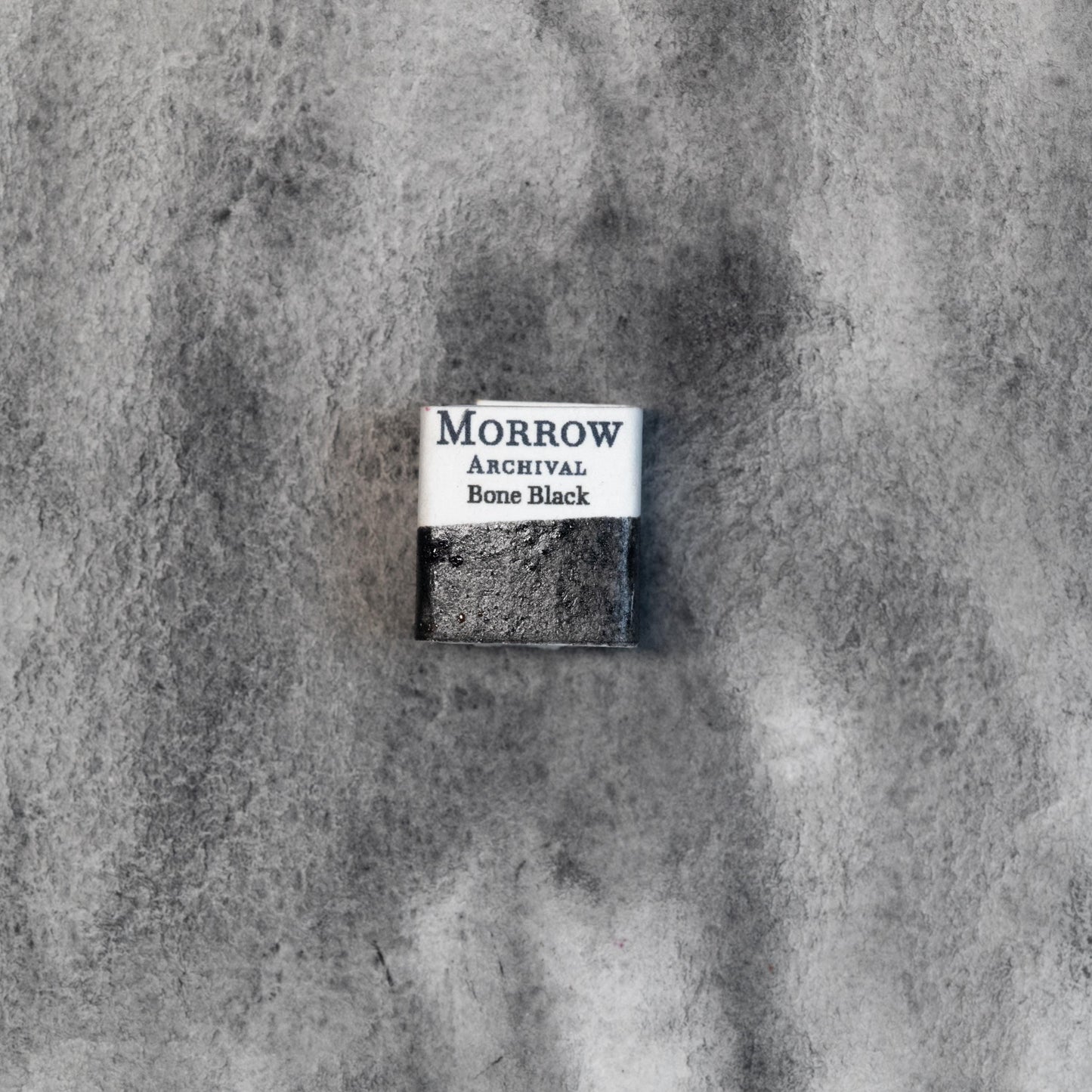 Close-up image of a half pan of Bone Black handmade watercolor paint, featuring a rich, matte black hue with a slightly warm undertone and natural granulation. The pan rests on a neutral background, highlighting its historic animal-derived origin and artisanal craftsmanship. Ideal for artists seeking a deep, natural black with classical texture and tonal richness.