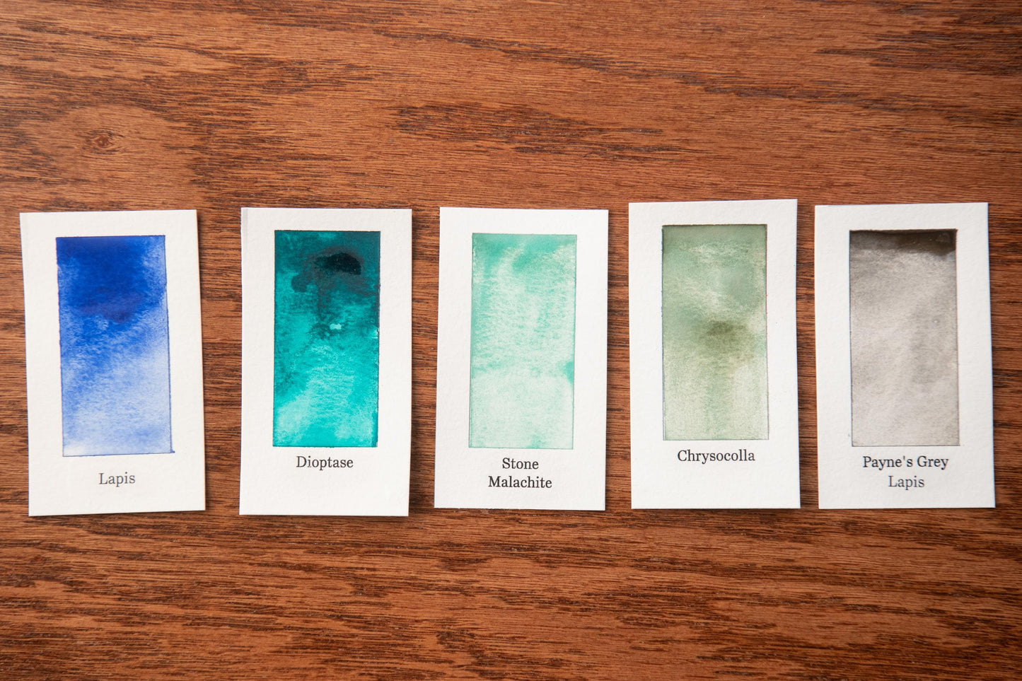 five watercolor paintings with different color variations, arranged on a wooden surface.
