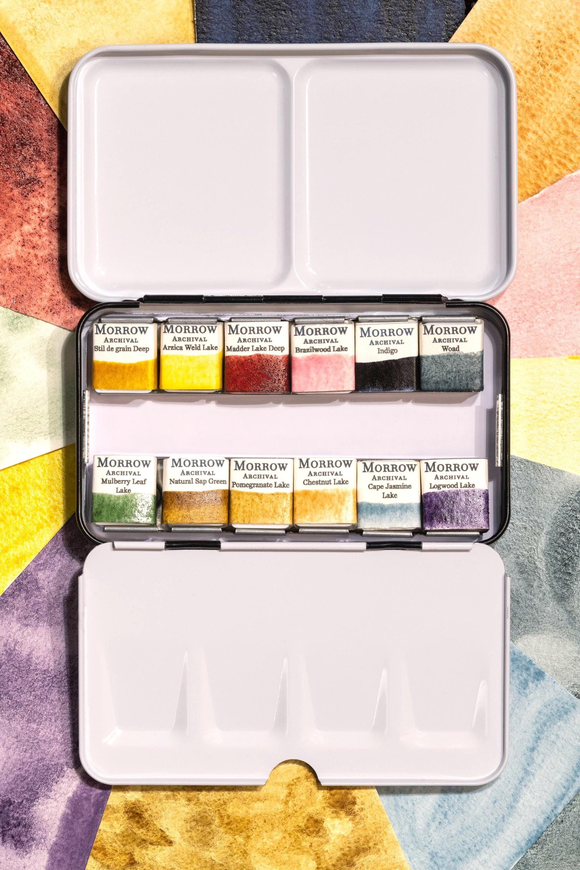 Archival watercolor palette set with 12 handmade pans labeled elegantly, shown with painted swatches behind. Includes Stil de Grain Deep, Arzica Light, Madder Lake Deep, Brazilwood Lake, Indigo Pigment, Woad Pigment, Mulberry Leaf Lake, Natural Sap Green, Pomegranate Lake, Chestnut Lake, Cape Jasmine Lake, and Logwood Lake. Plant-based pigments crafted with honey and gum arabic, ideal for historical palettes and botanical work.