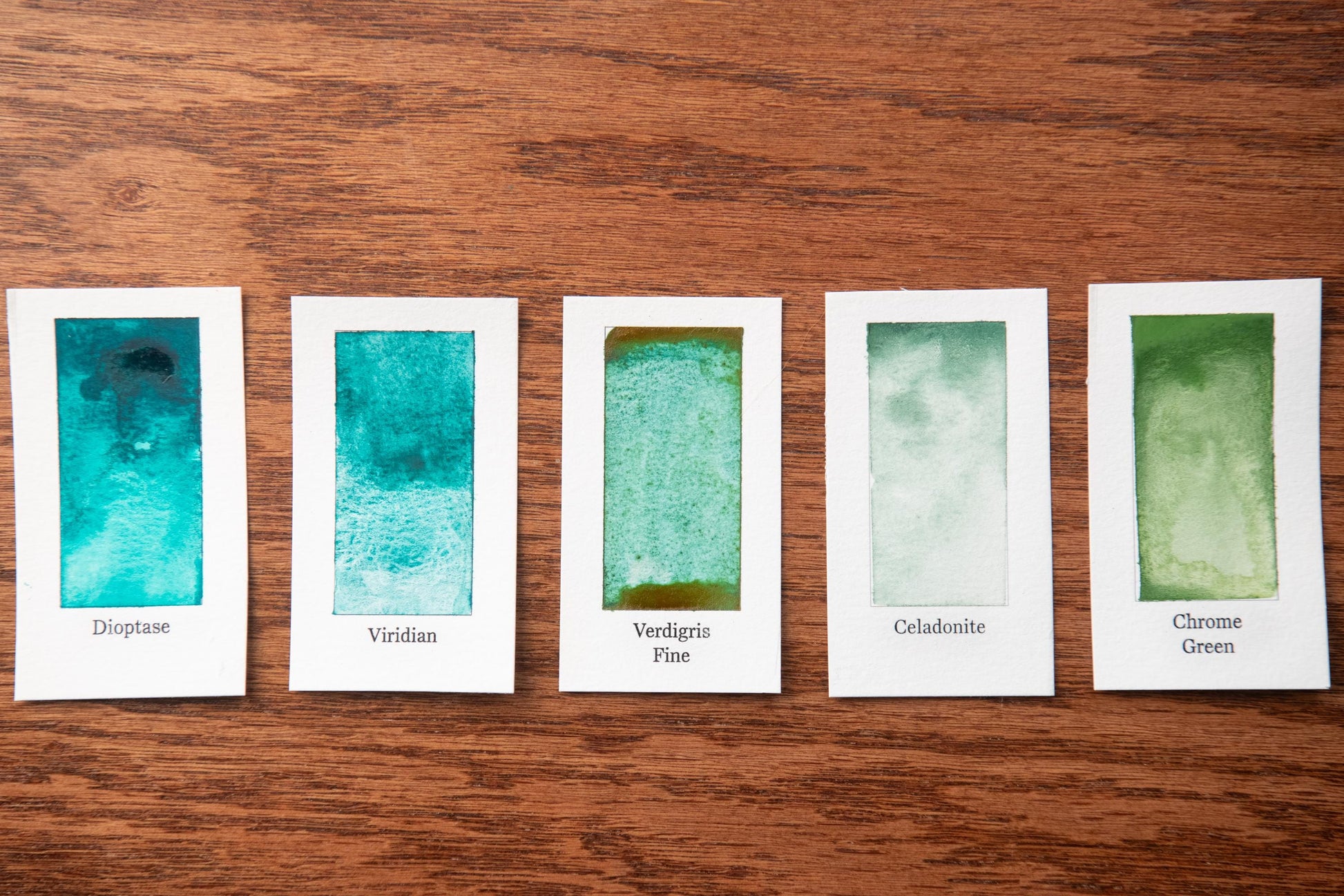 five small white cards with different shades of green paint, arranged in a row on a wooden surface.