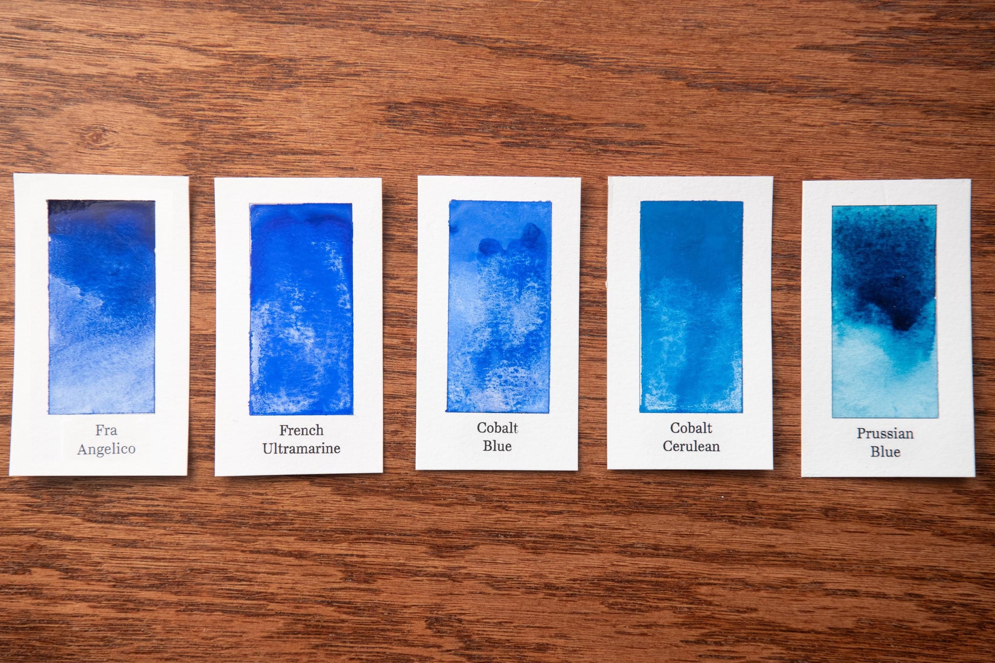 five watercolor paintings in various shades of blue, arranged on a wooden surface.