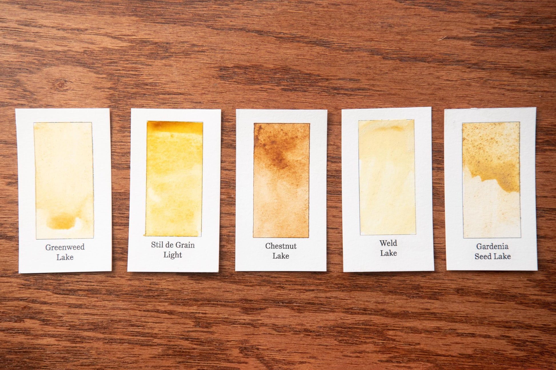 five watercolor paintings on a wooden surface, each with a different color and texture, arranged in a row.