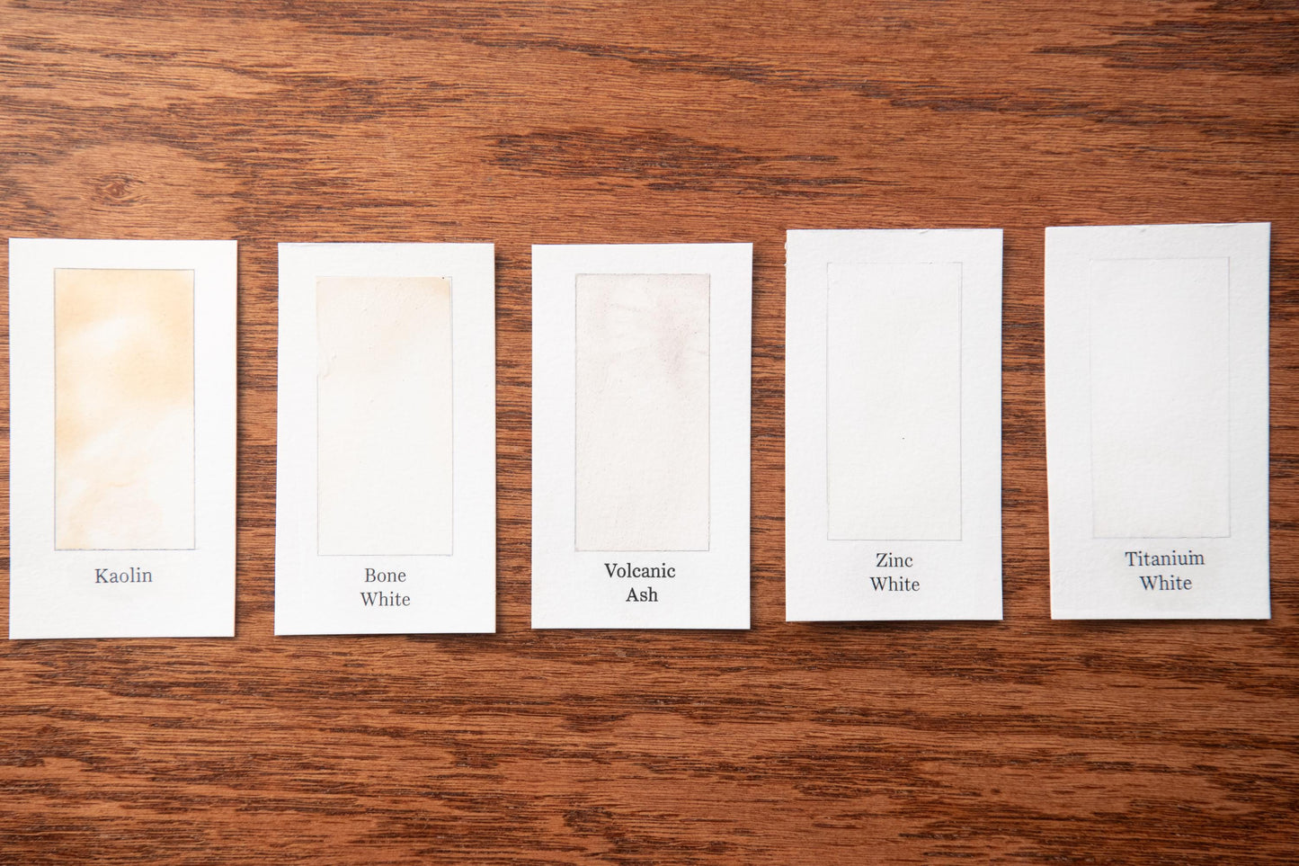 five white cards with different paint colors labeled on them, arranged on a wooden surface.