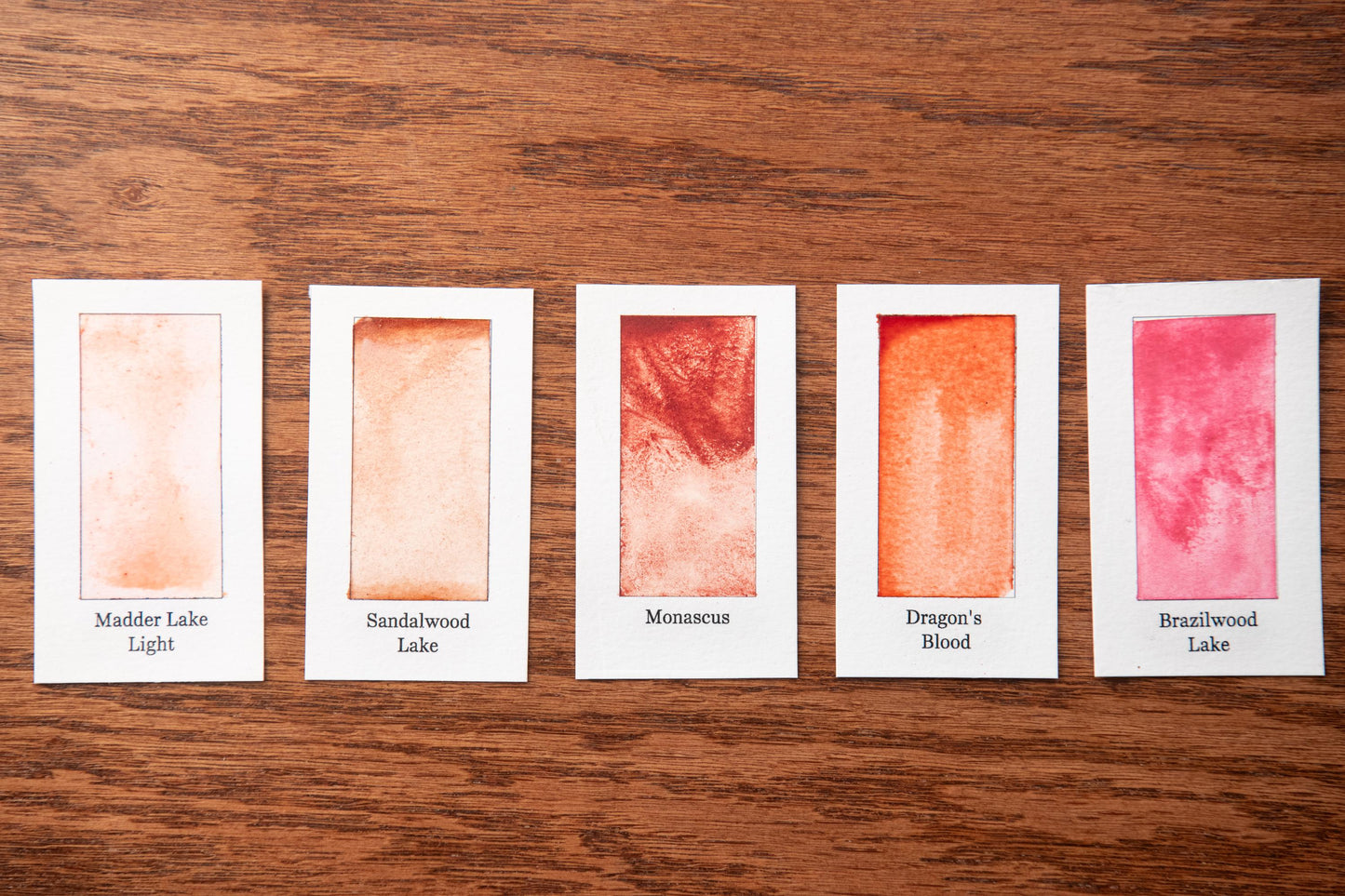 five small white cards with different colored paint swatches on them, arranged on a wooden surface.
