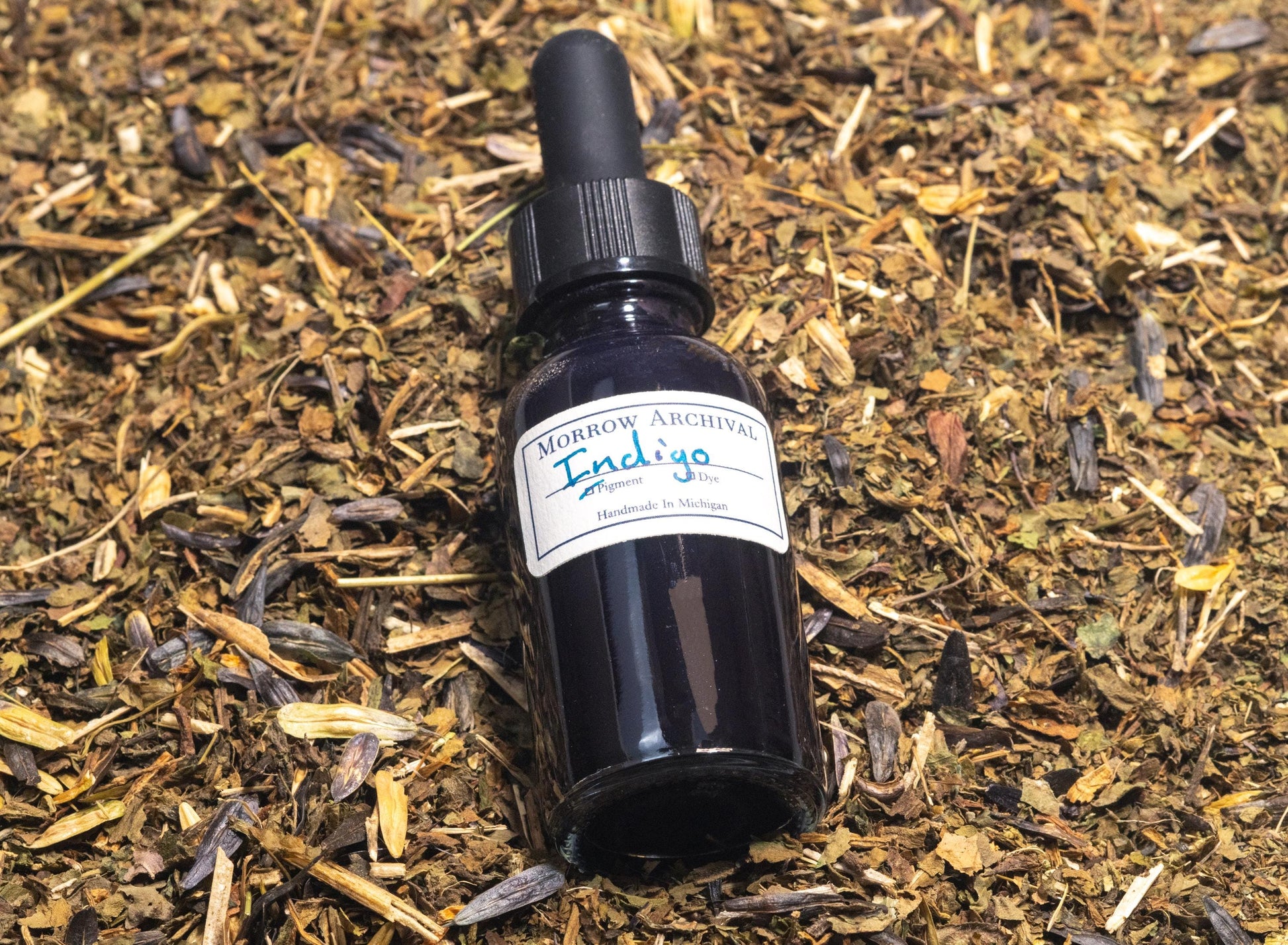 Glass bottle of natural Indigo ink resting on dried Indigofera tinctoria leaves and indigo cakes. The scene connects the finished ink to its botanical source, revealing the deep blue dye‚Äôs rich cultural and historical roots.