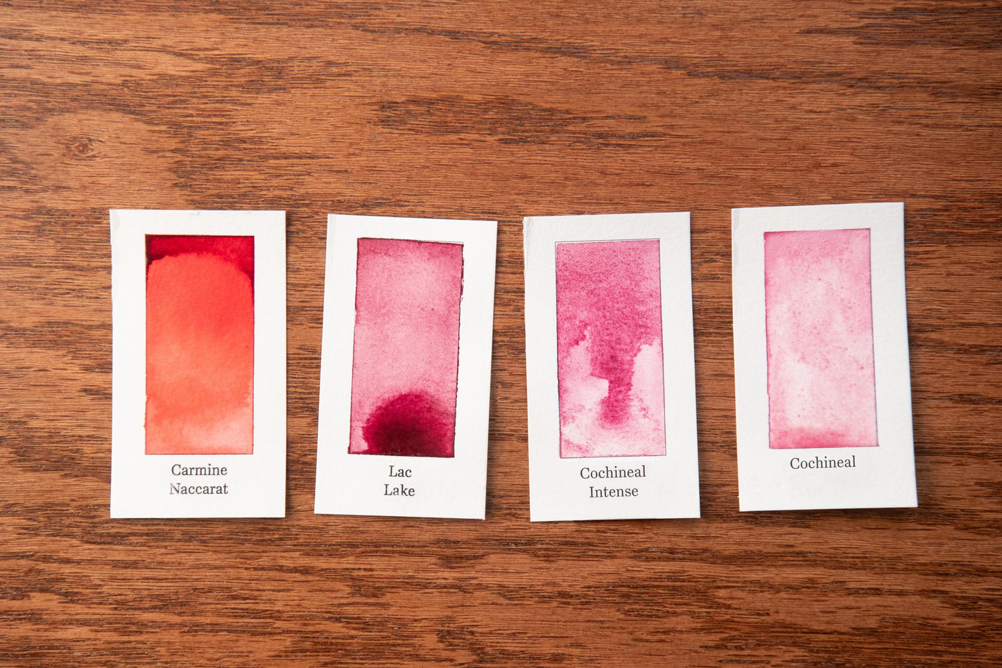 four small white cards with different watercolor paint swatches on a wooden surface.